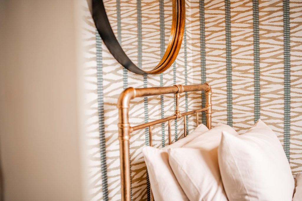 A close-up shot from a WildKind Interiors-styled bedroom in Anfield, Liverpool, showcasing a striking brass bedframe set against bespoke neutral and teal striped wallpaper. The image...