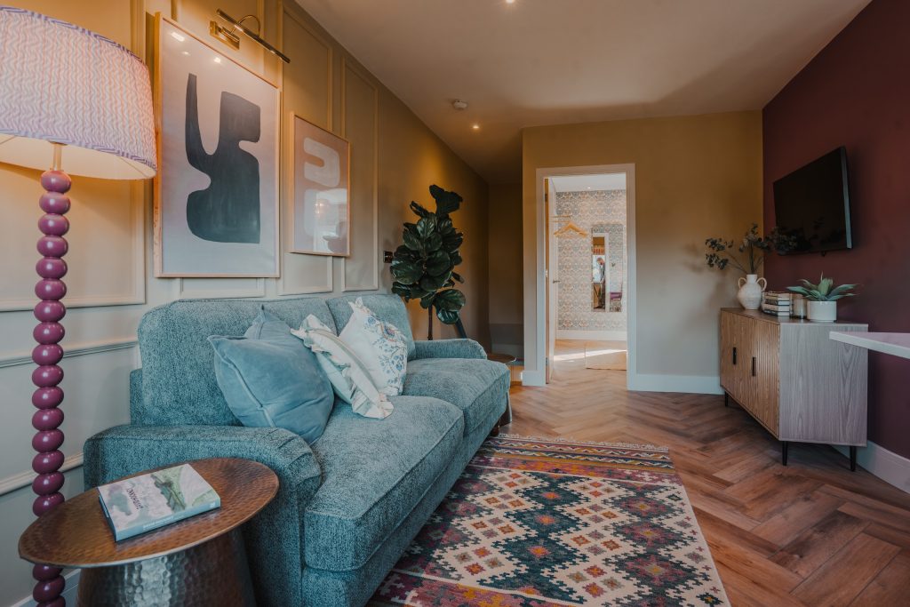 Explore this chic sitting room in Beckwithshaw, North Yorkshire serviced accommodation. Featuring a plush teal sofa with patterned and solid cushions against a paneled feature wall, a tall...
