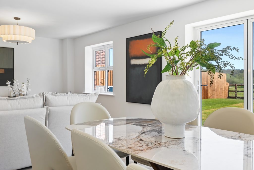Open-plan dining area with a marble-effect table, cream chairs, a sculptural vase of greenery and a leafy garden view.