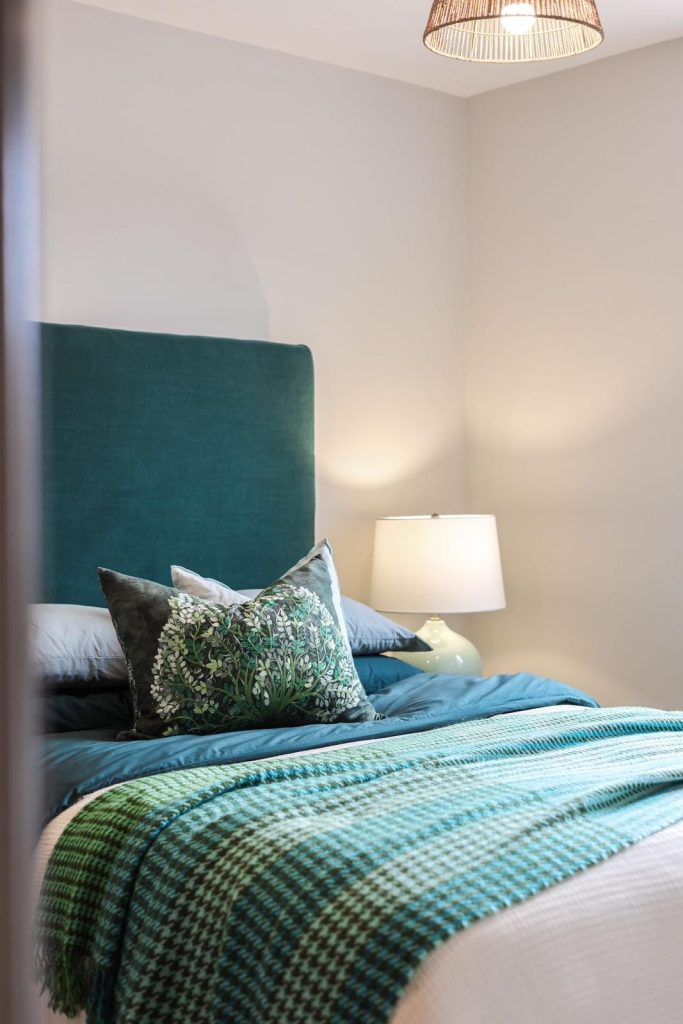 A stylish bedroom staged by WildKind Interiors in Huntingdon, York. Highlighted by a teal upholstered headboard, layered bedding in coordinating blues, a patterned green and black accent...