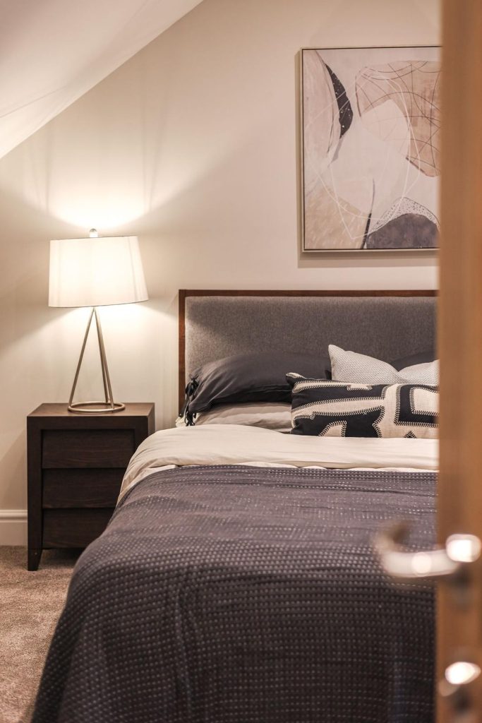 An inviting attic-style bedroom staged by WildKind Interiors in Huntingdon, York. This scene features a grey upholstered bed dressed in layered bedding, a dark wood bedside table...