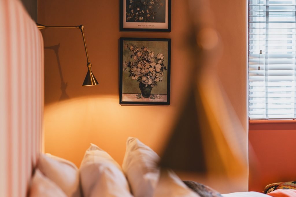 Bedroom detail with a striped headboard, a brass wall light and a framed floral print in warm evening light.