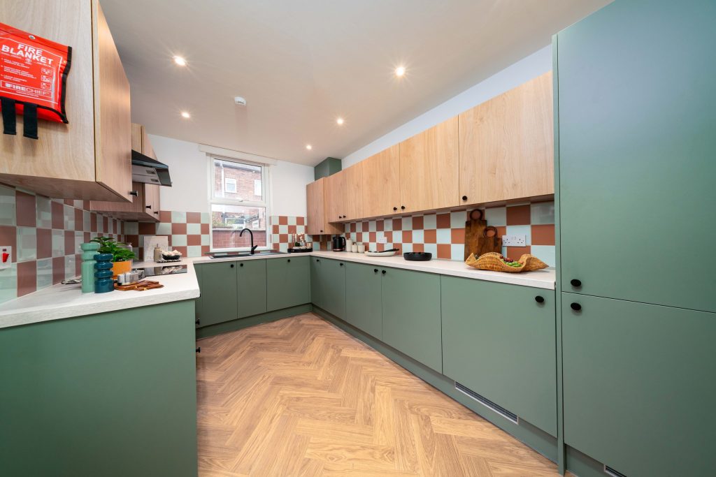 Communal kitchen with sage-green cabinetry, oak uppers, a checkerboard splashback and herringbone flooring.