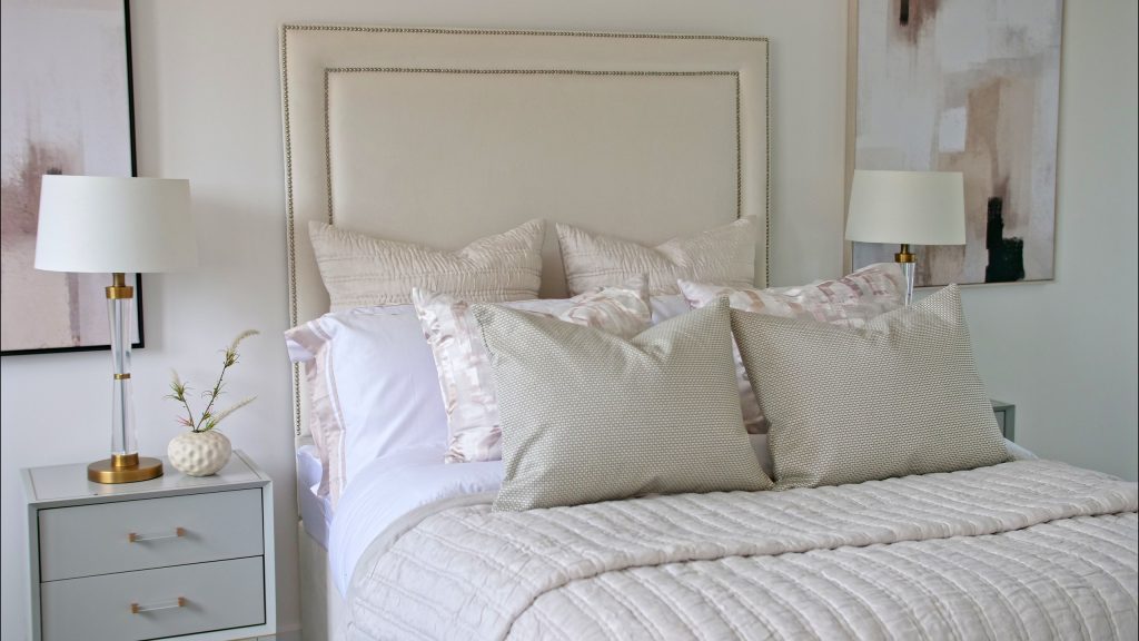Bedroom styled with an upholstered headboard, layered cream cushions and matching bedside lamps beneath soft abstract artwork.