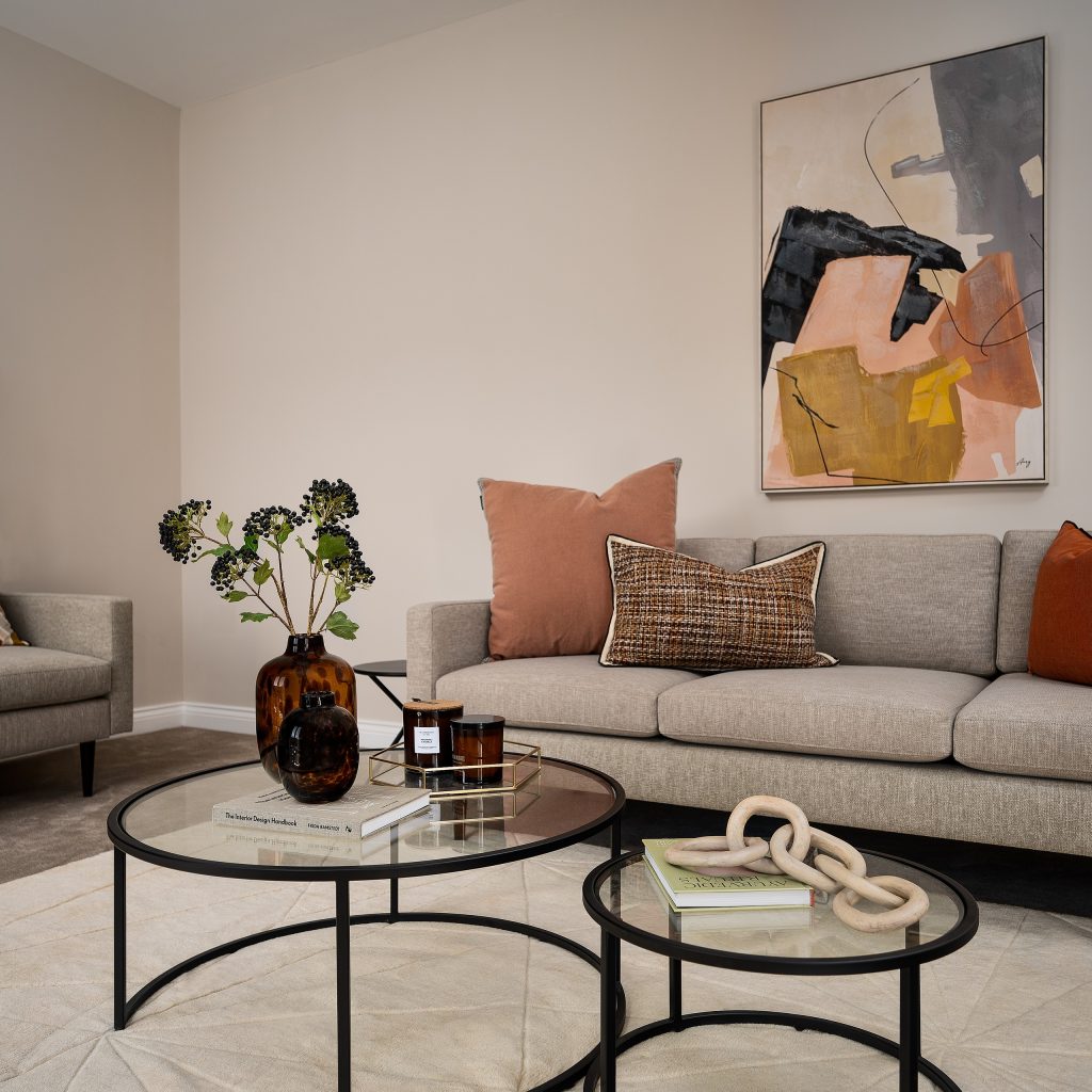 A warm living room scene staged by WildKind Interiors, featuring a neutral-toned sofa layered with rust and patterned accent pillows, nested glass coffee tables showcasing amber vases...