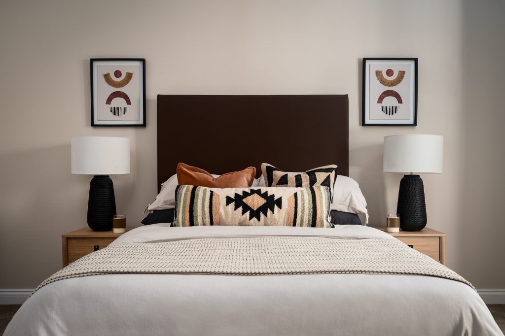 A modern bedroom expertly staged by WildKind Interiors, featuring a dark upholstered headboard, layered decorative pillows with geometric patterns, matching bedside lamps on wooden...