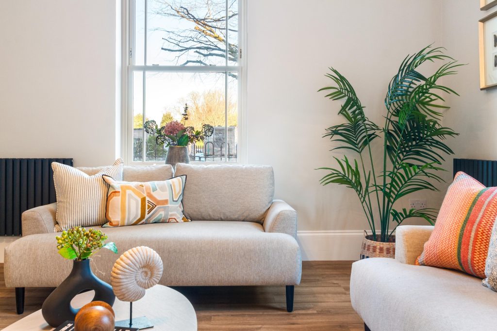 Living room with cream sofas, a round coffee table styled with a shell and sculptural decor, and a tall palm by the bright window.