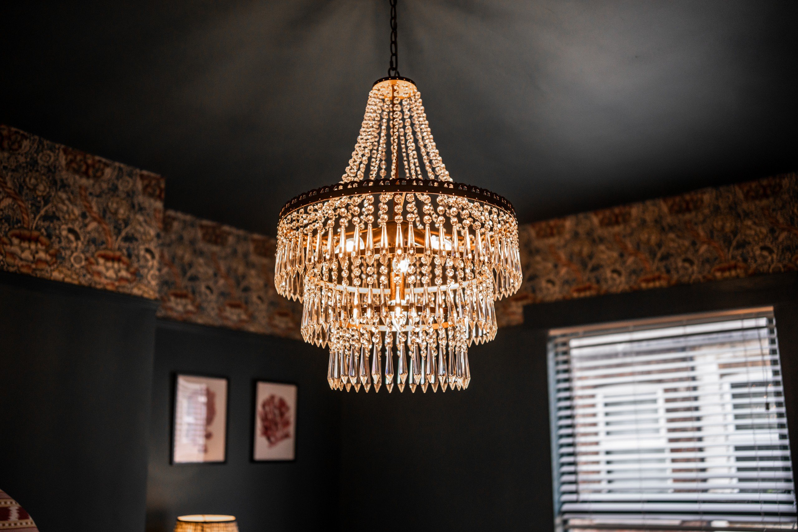 Elegant crystal chandelier in a room with dark walls and patterned wallpaper. Art frames and a window with blinds in the background.