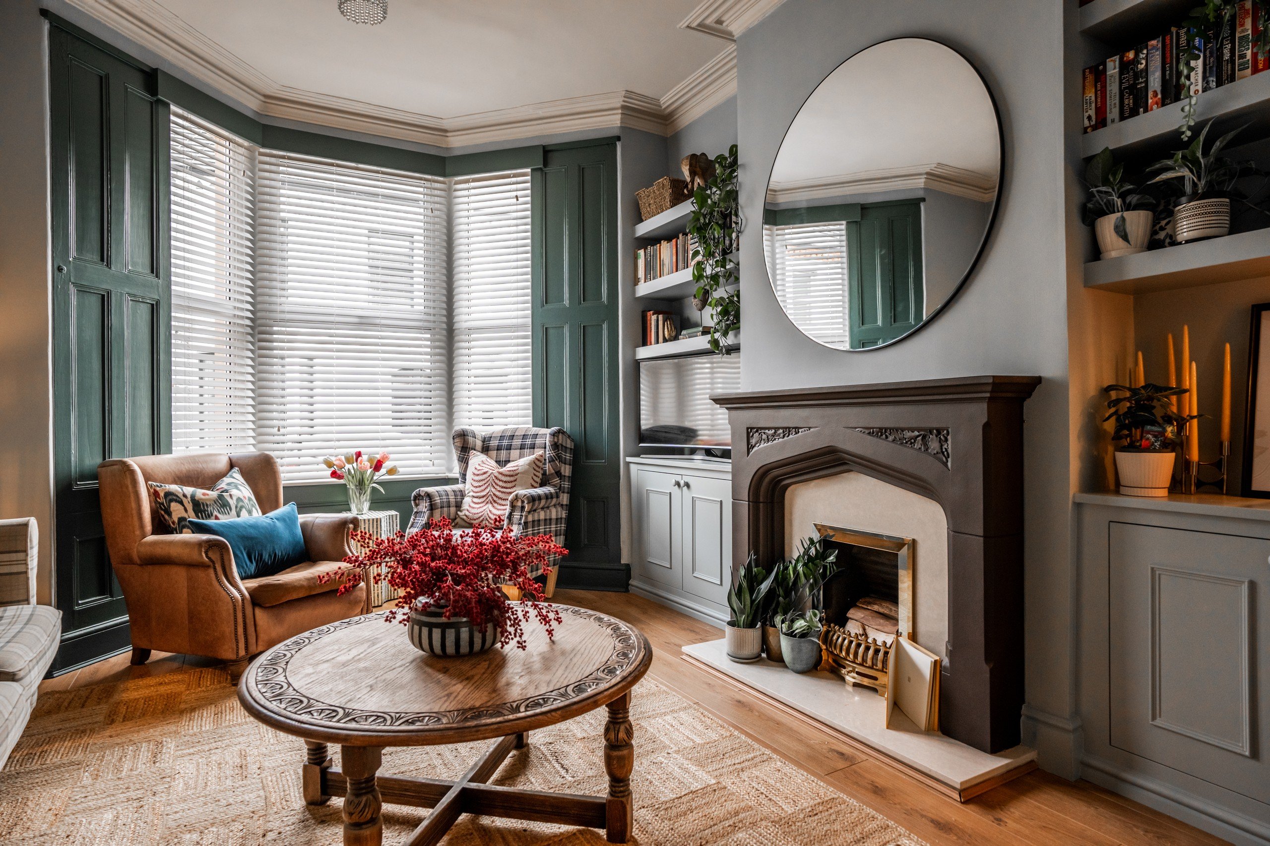 Cozy living room with green walls, brown armchair, plaid chair, round wooden table, red flowers, mirror above fireplace, bookshelves.