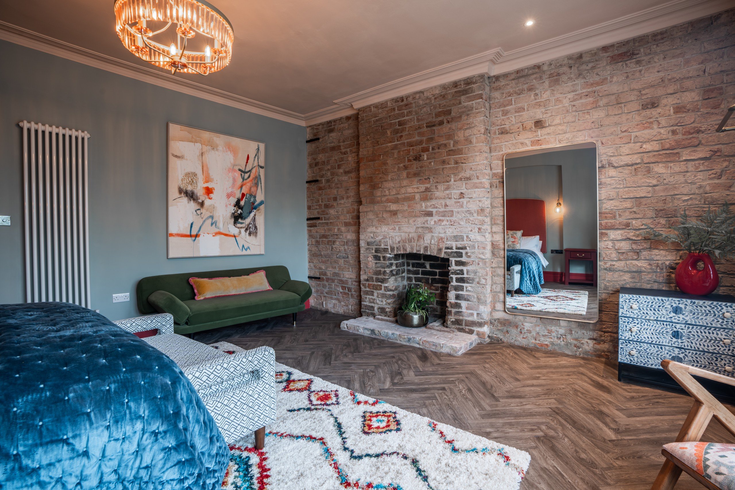 Cozy room with a green sofa, abstract painting, brick fireplace, and large mirror. Blue bedspread and patterned rug add color and texture.