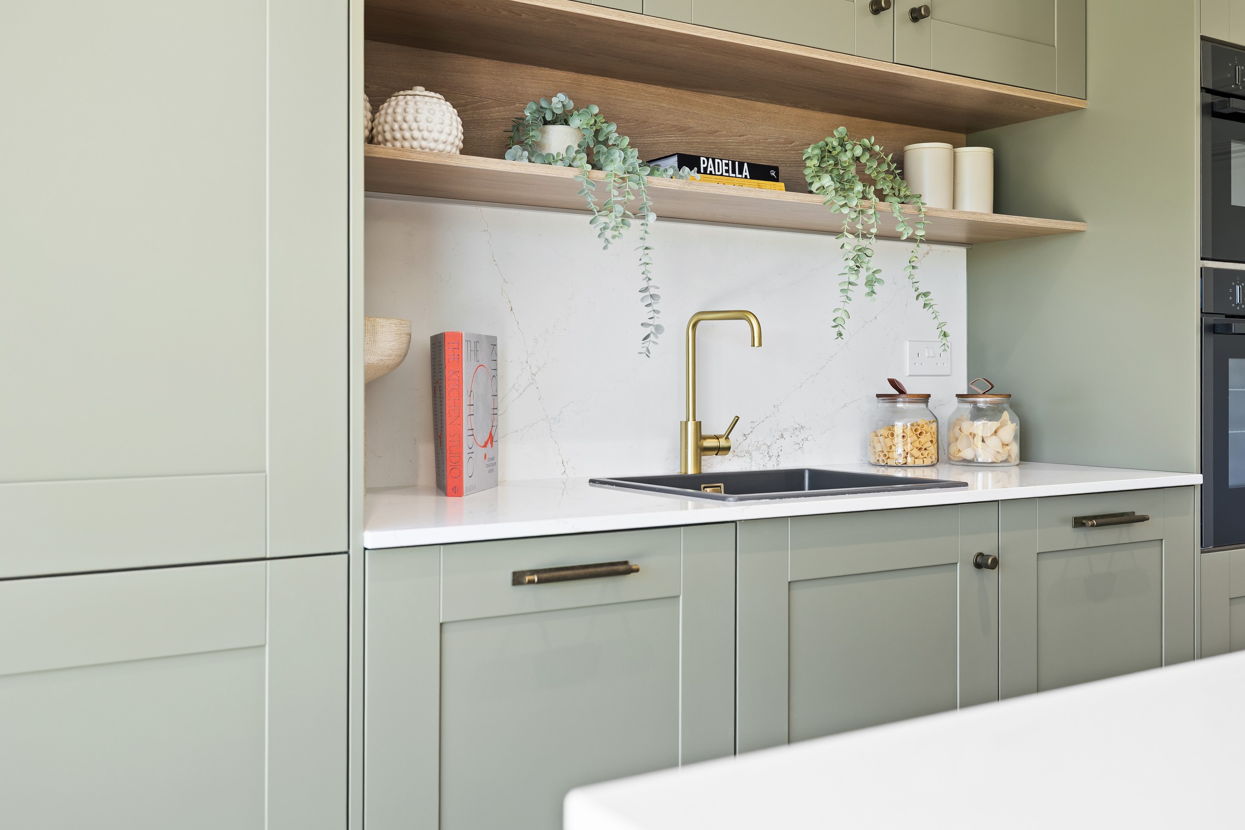 Modern sage-green kitchen with brass faucet, open shelves, trailing plants, pasta jars, and a PADELLA book above the sink.