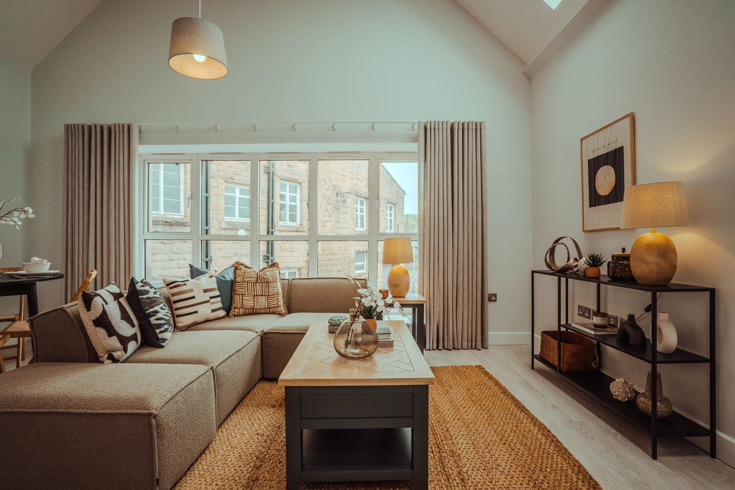 Cozy living room with a beige sofa, patterned cushions, coffee table, and potted plant. Large window, neutral tones, and warm lighting.