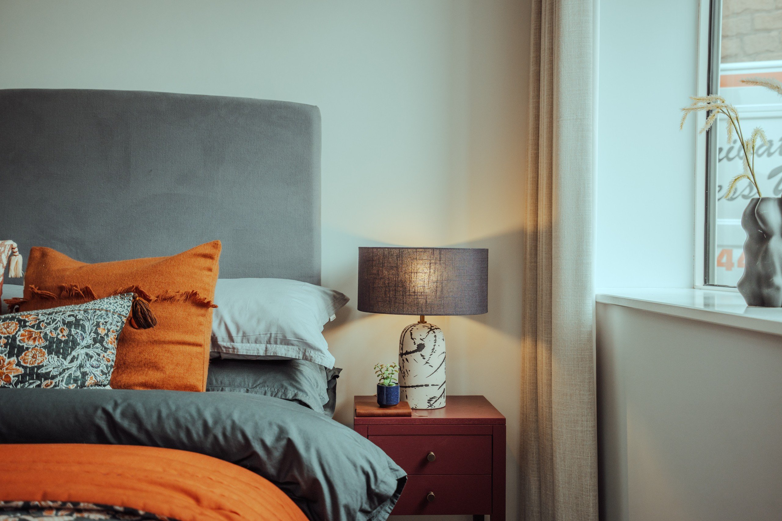 Cozy bedroom with a gray headboard, orange and floral pillows, a red nightstand, and a lit lamp. Window with soft light and sheer curtains.