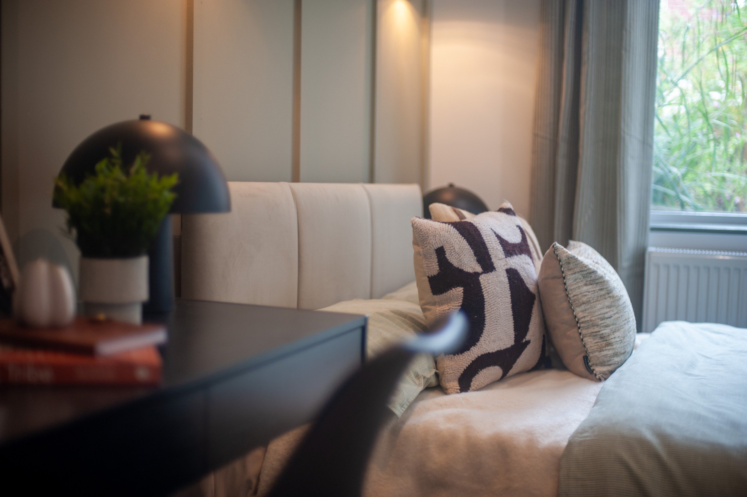 Cozy bedroom with a bed adorned with patterned pillows, a sleek lamp, and books on a side table. Soft lighting and window view.