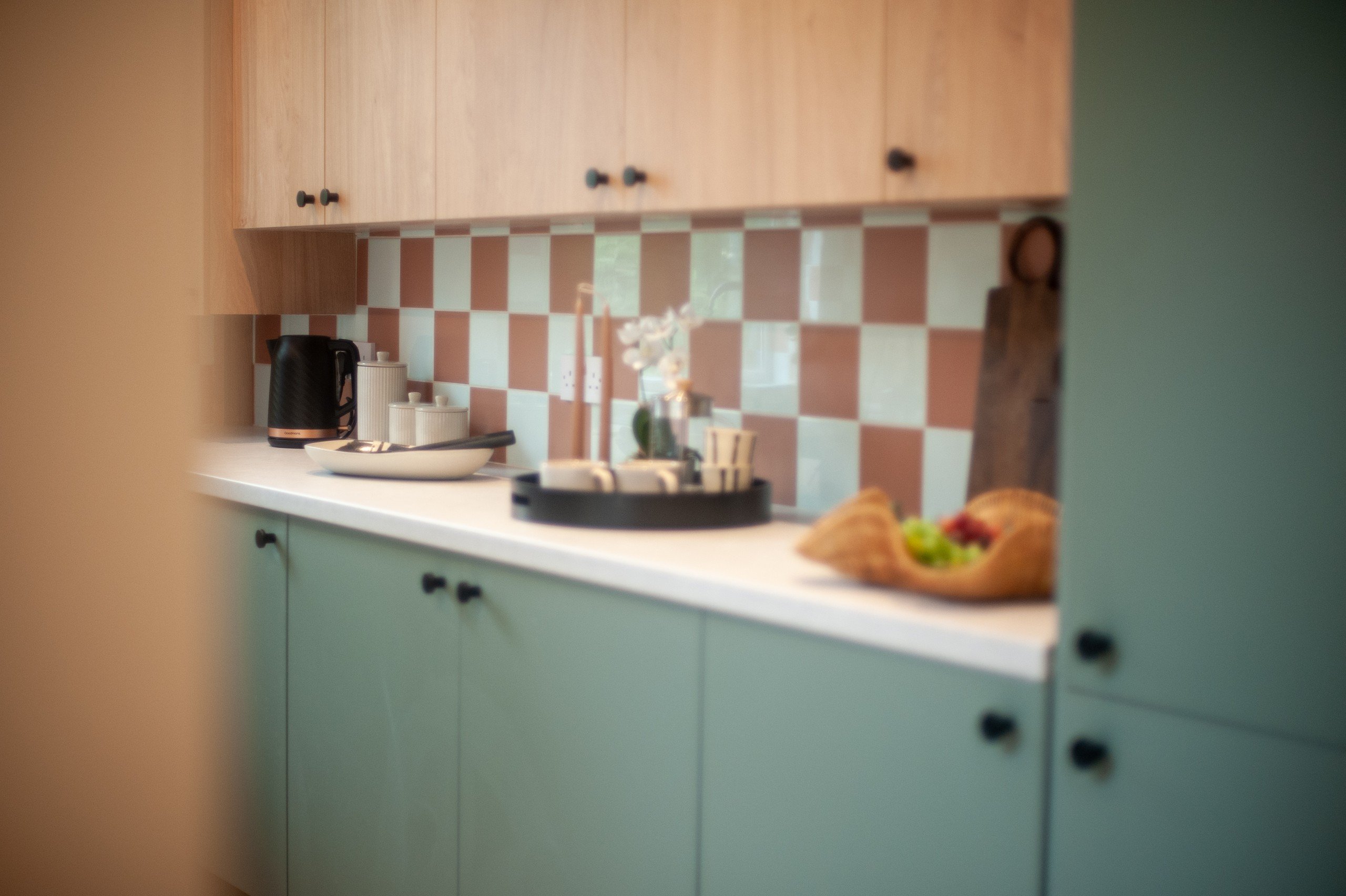 Modern kitchen with a checkered backsplash, wooden cabinets, and a countertop holding a kettle, tray with cups, and a fruit basket.