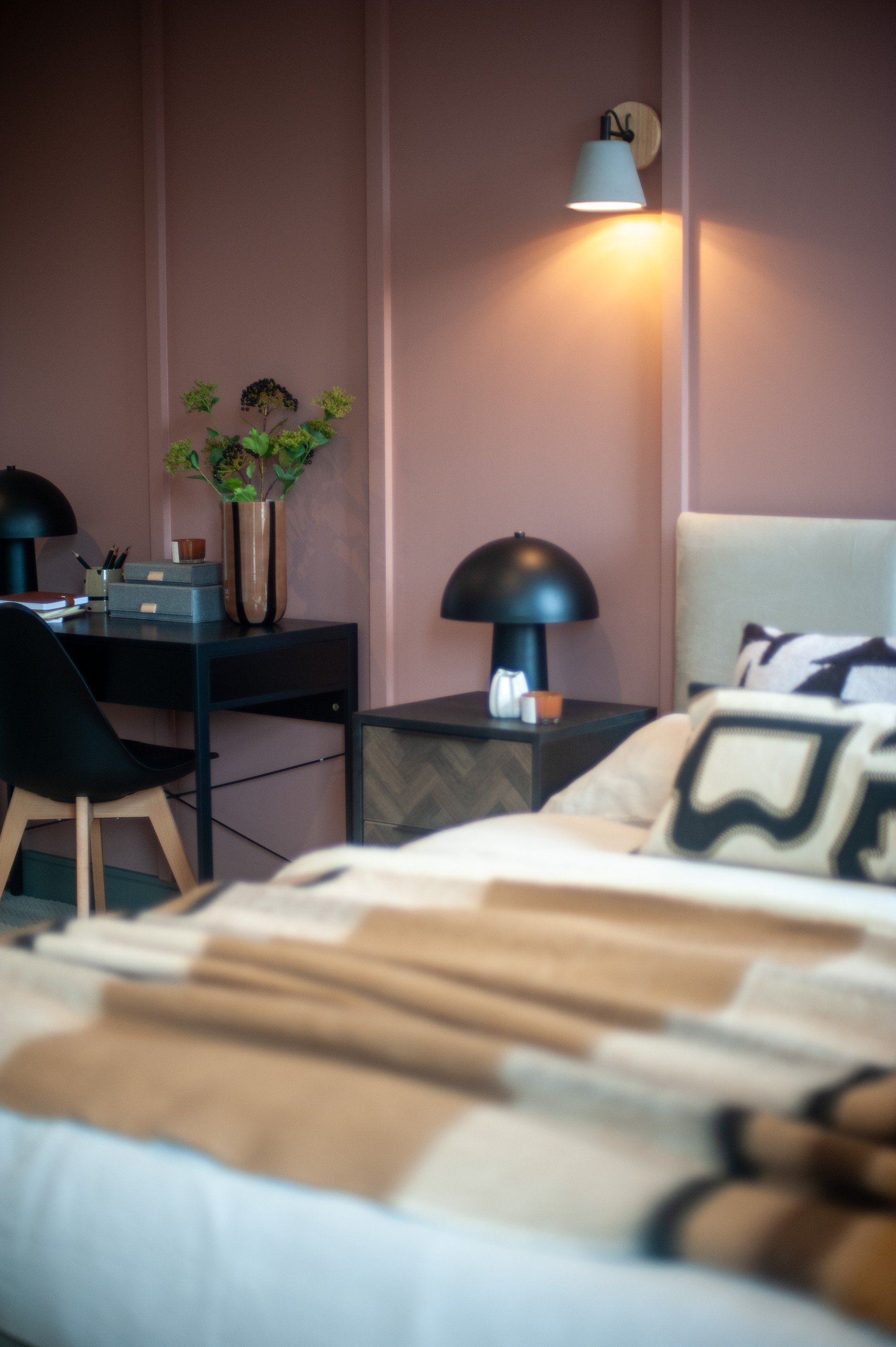 Cozy bedroom with soft pink walls, a bed covered with a beige blanket, a desk with black lamps, plants, and a lit wall sconce.