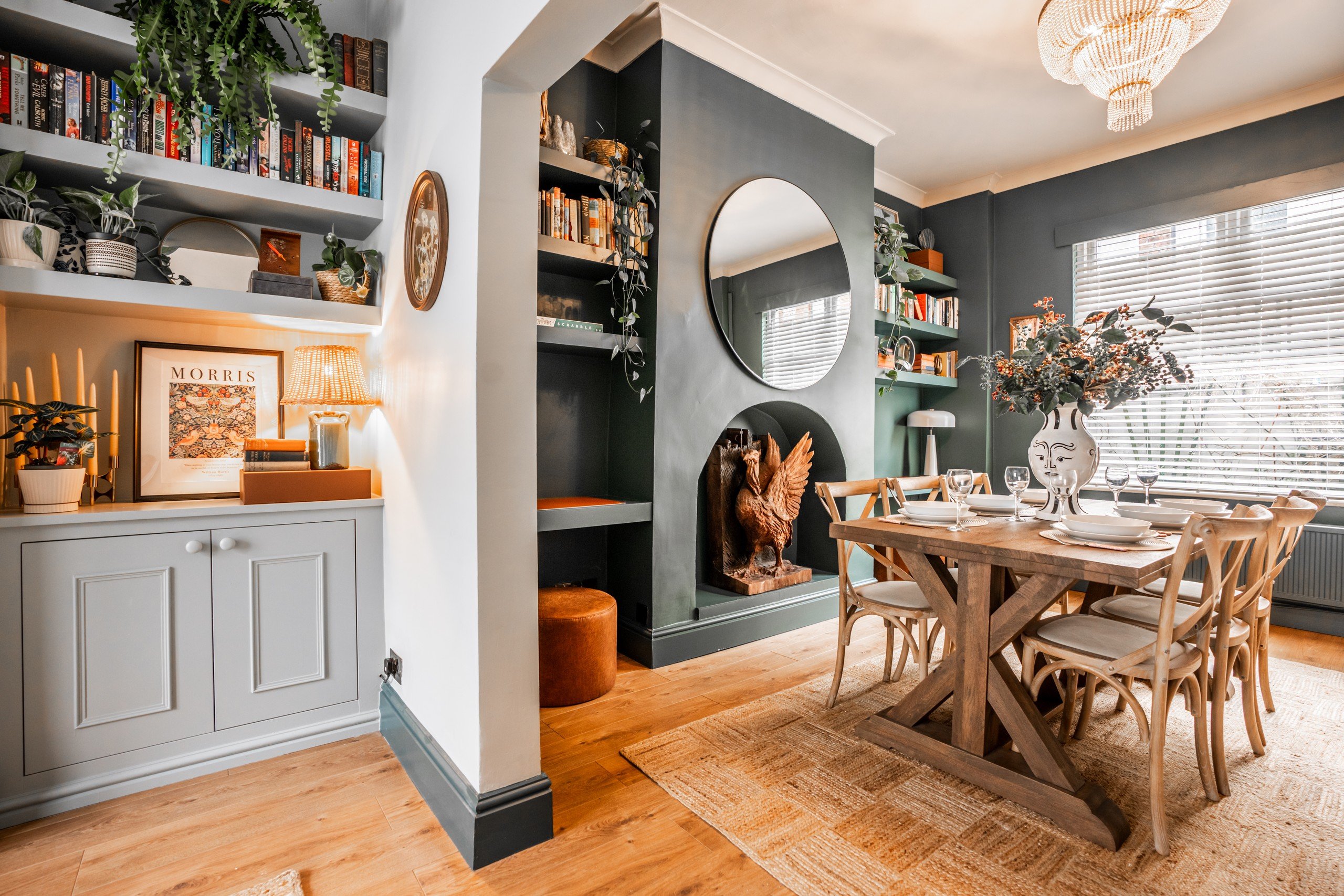 Cozy dining room with a wooden table set for guests. Decor includes plants, books, a large mirror, art, and warm lighting.