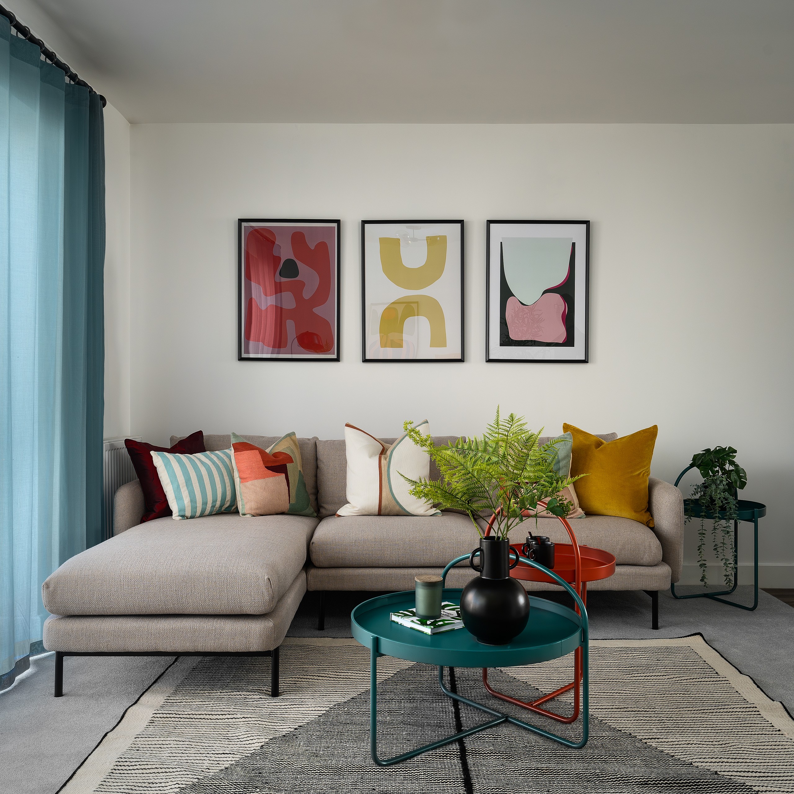 Stylish living room with beige sectional, colorful pillows, abstract wall art, teal curtains, and nested teal-orange coffee tables.