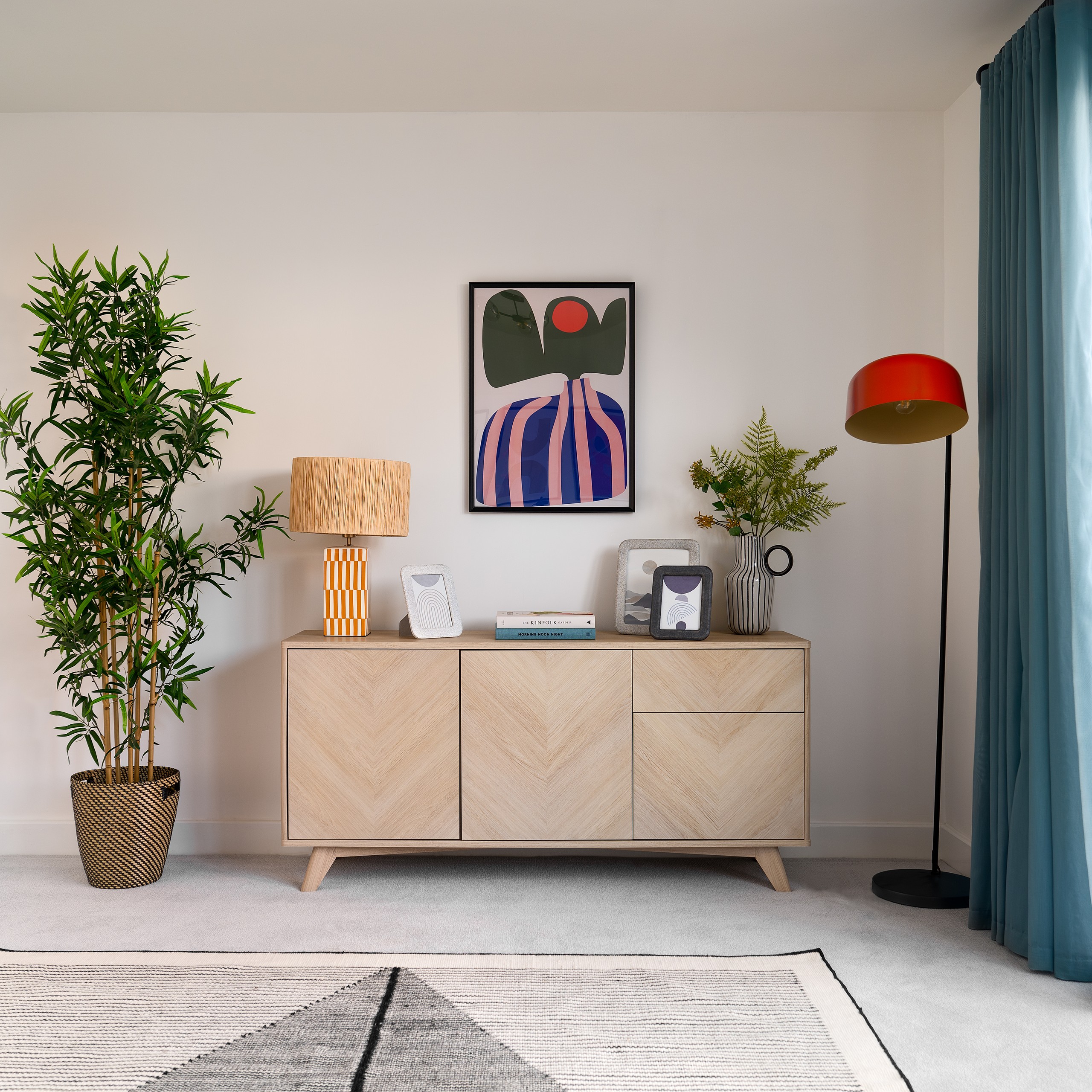 Modern living room with wood sideboard, abstract wall art, plants, colorful lamps, blue curtains, and Kinfolk books on display.