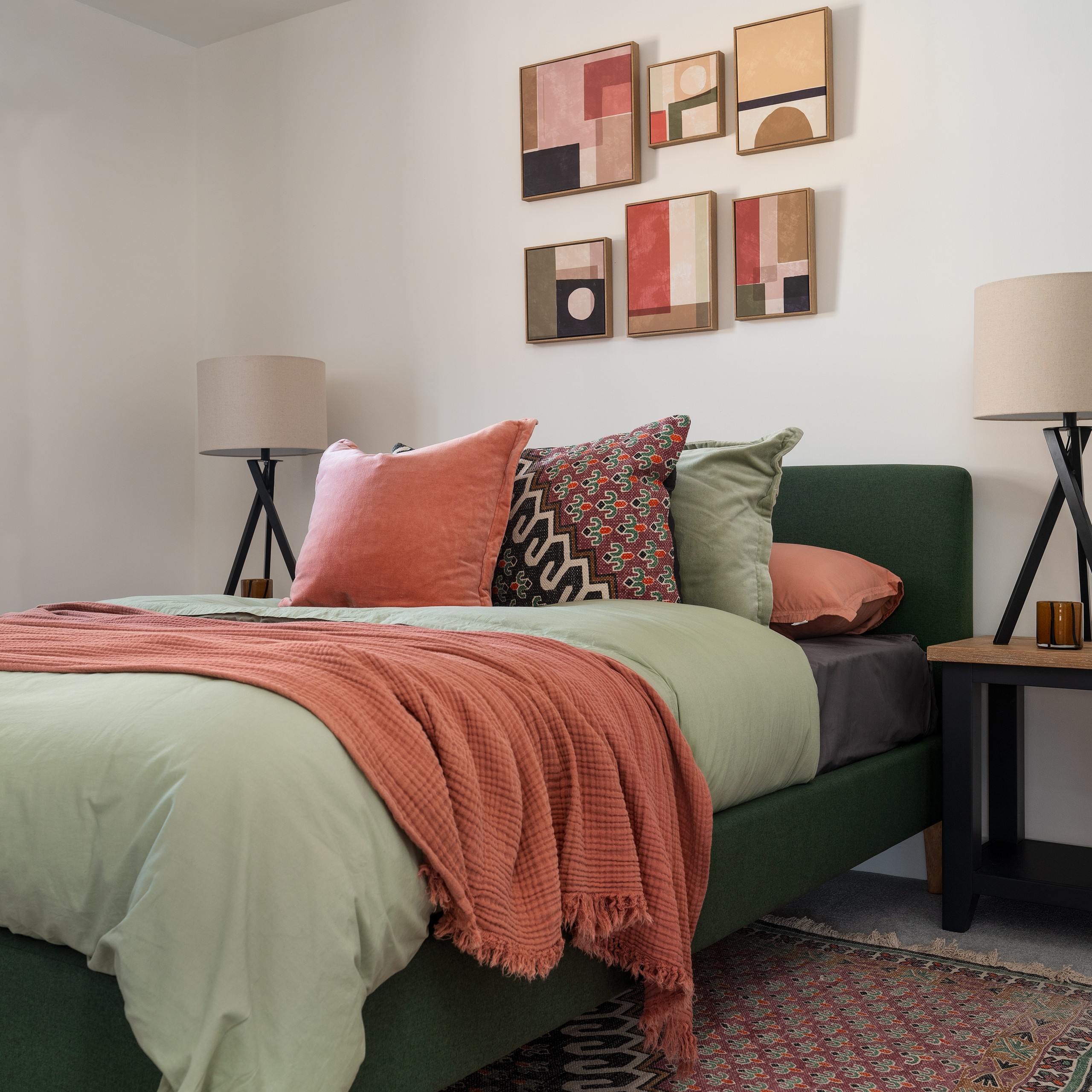Cozy bedroom with green bed, pastel bedding and patterned pillows, flanked by lamps and abstract wall art on a white wall.