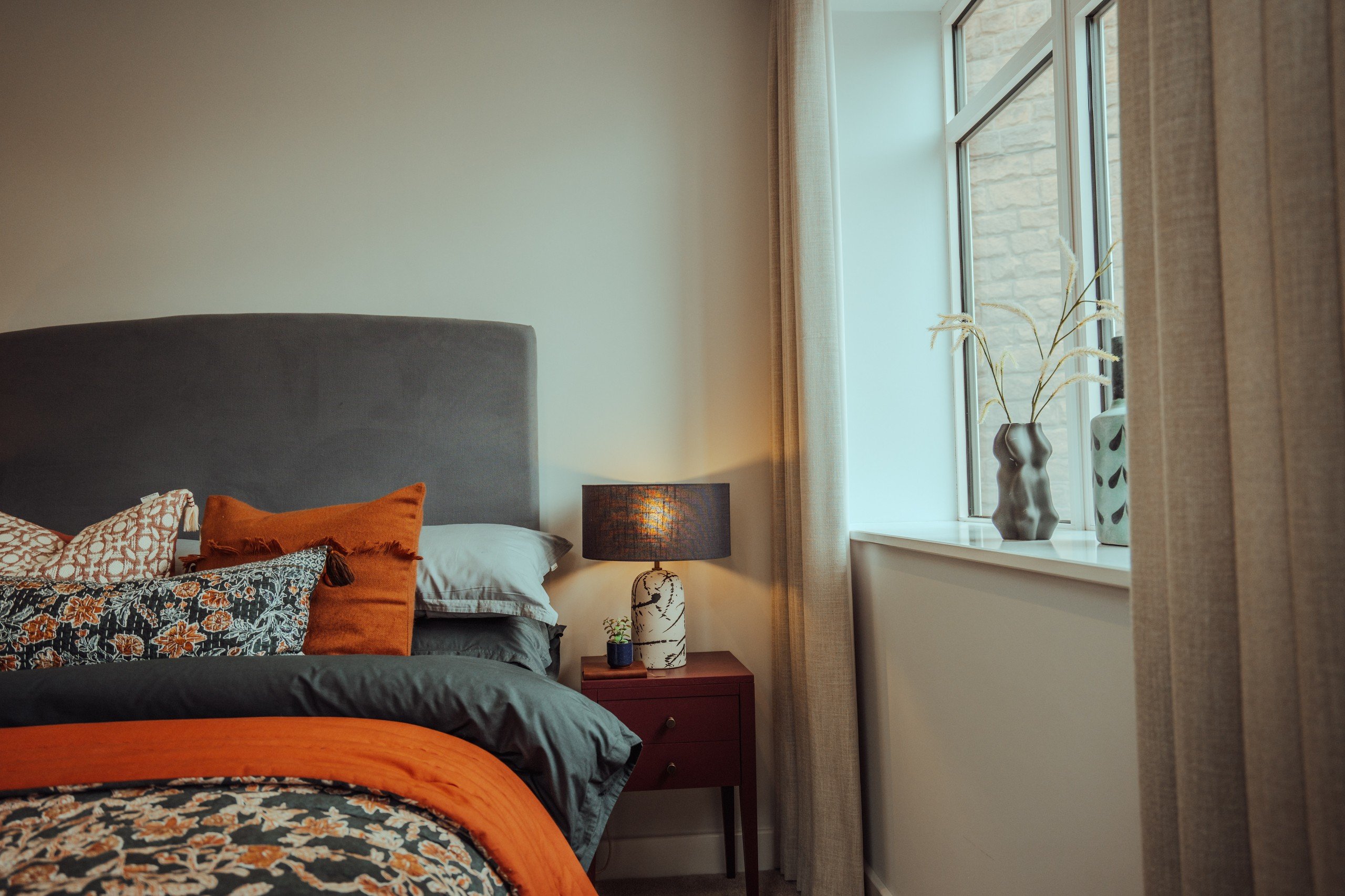 Cozy bedroom with a gray headboard, colorful pillows, orange accents, and a floral bedspread. A lamp on a bedside table and a window with vases.