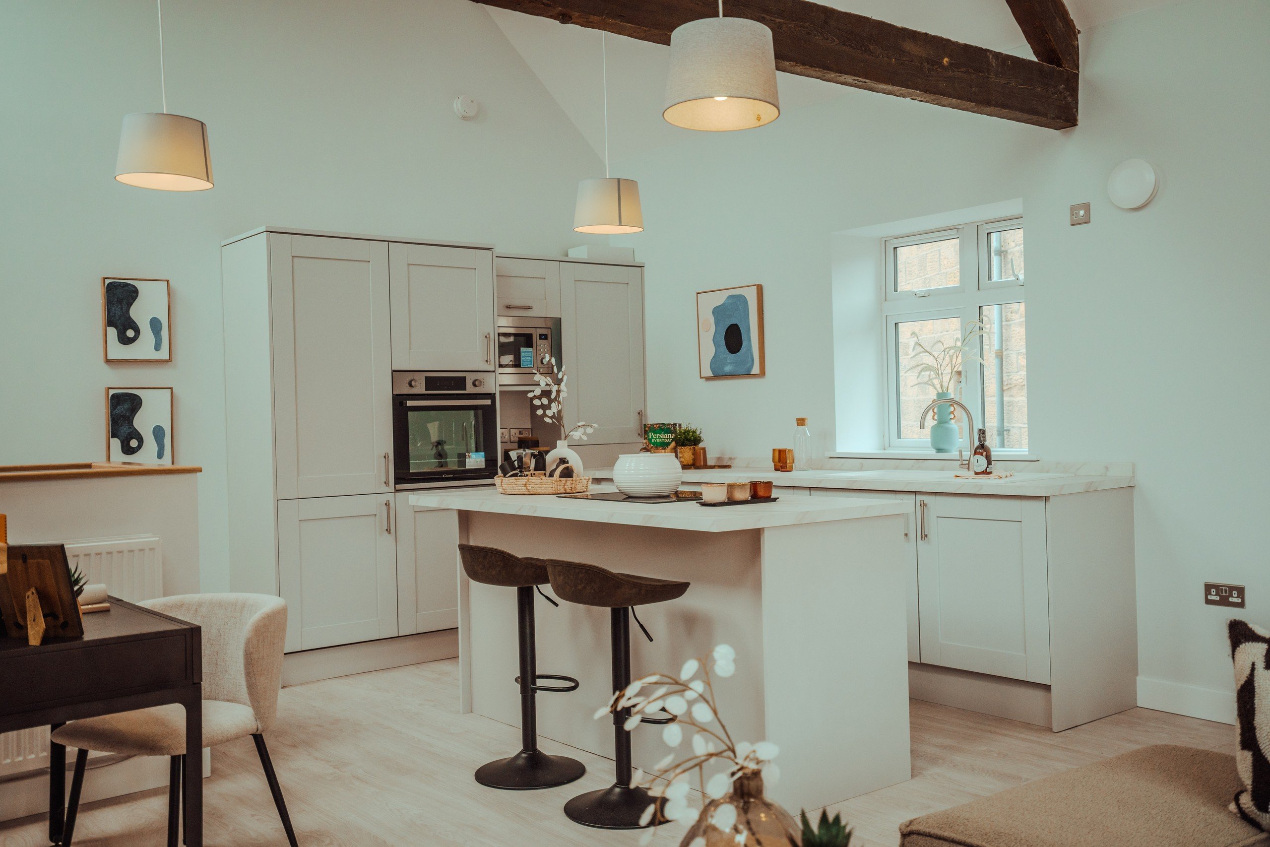 Modern kitchen with a white island, two bar stools, and wooden beams. Abstract art on walls, potted plants, and warm lighting create a cozy feel.