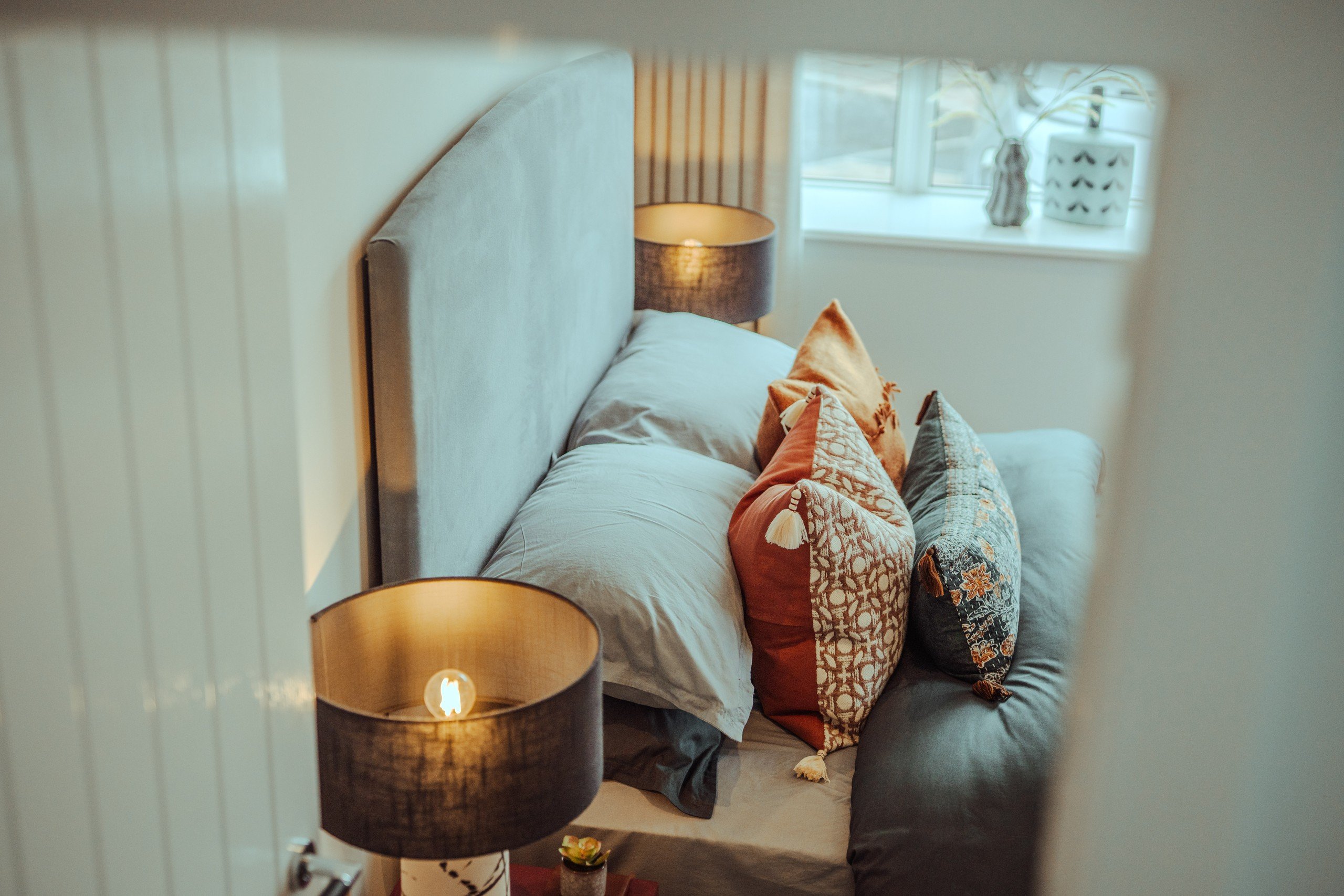 Cozy bedroom with a gray bed, colorful patterned pillows, and lit lamps. A window with decor creates a warm, inviting atmosphere.