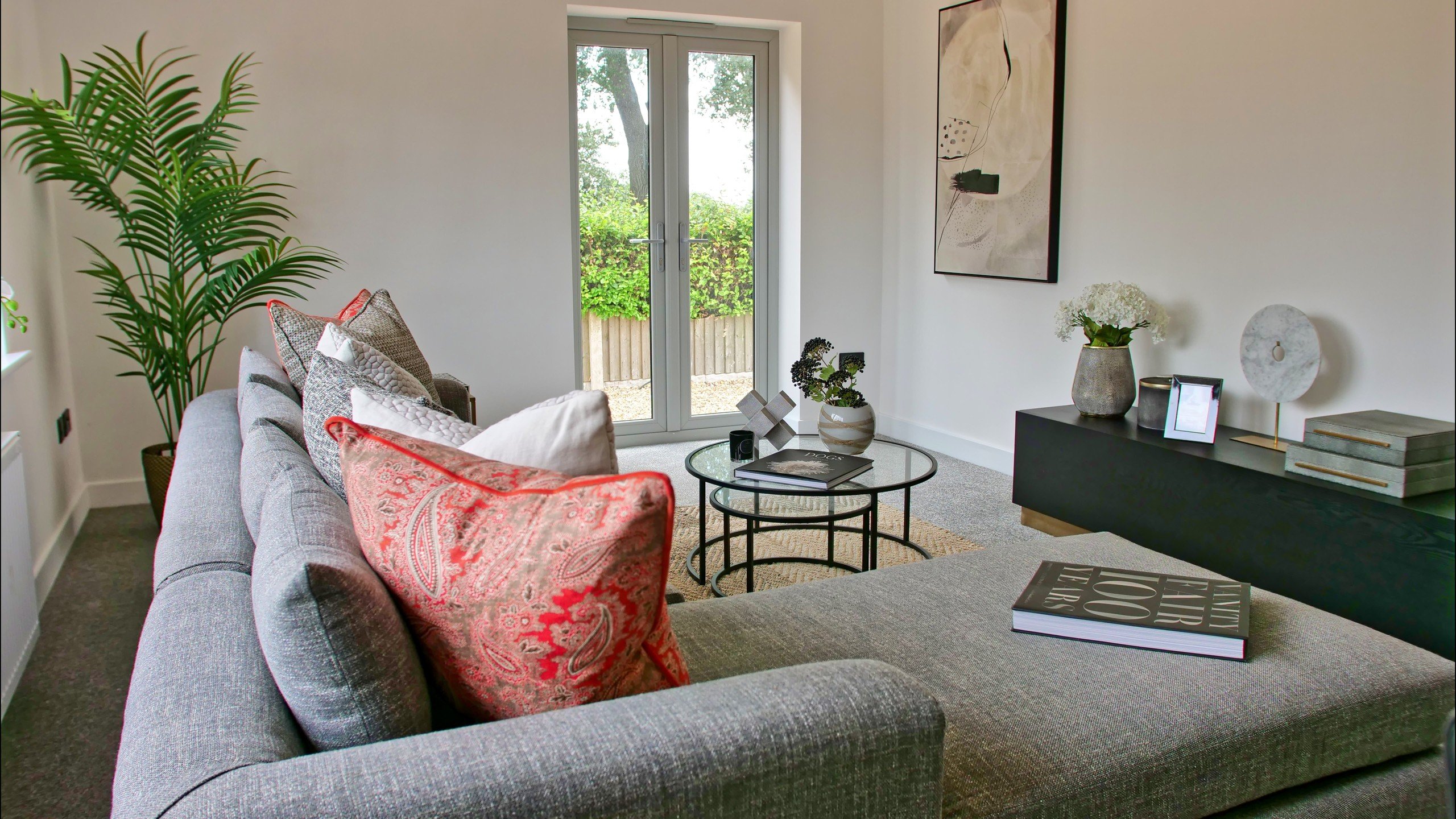 WildKind designed. Modern living room with a gray sofa, colorful pillows, glass coffee table, and a large plant. Art on walls, light from glass doors. Cozy vibe.