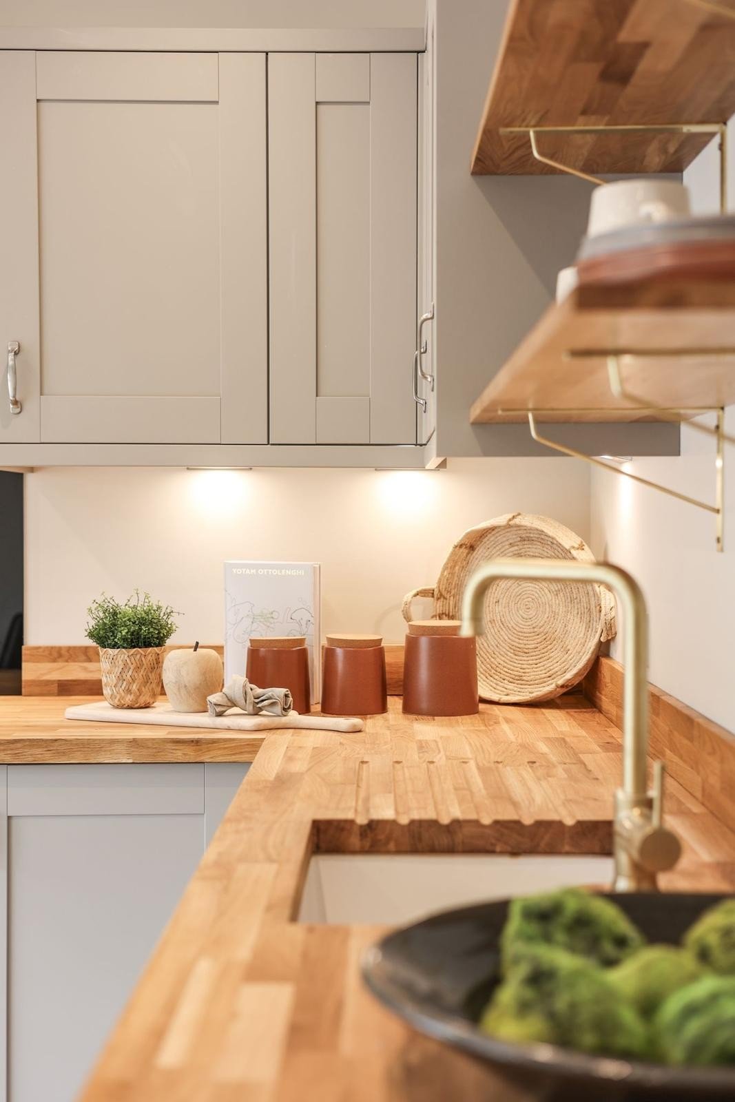 WildKind Designed modern kitchen with wooden countertops, tan cabinets, potted plant, canisters, cookbook, woven tray, and round gold faucet; warm lighting.