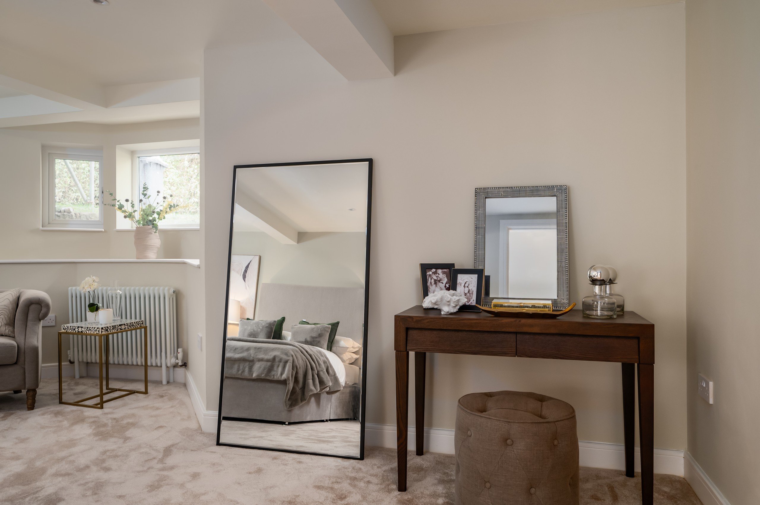 WildKind designed bedroom with neutral tones; a large mirror reflects a bed. Wooden vanity with framed photos and accessories.