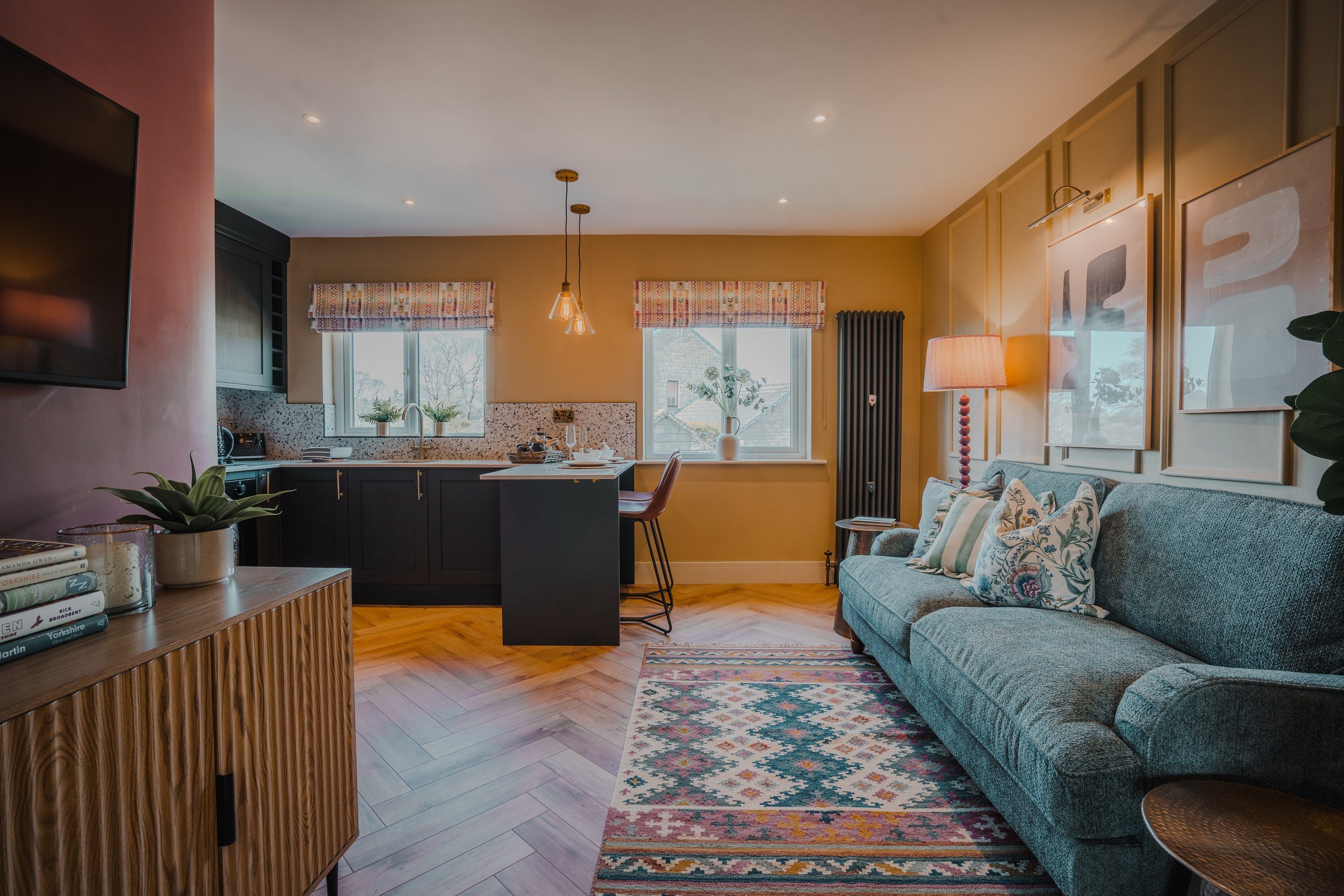Cozy living room with a blue sofa, colorful rug, and wooden cabinets. Pendant lights over kitchen island. Warm lighting and decorative pillows.