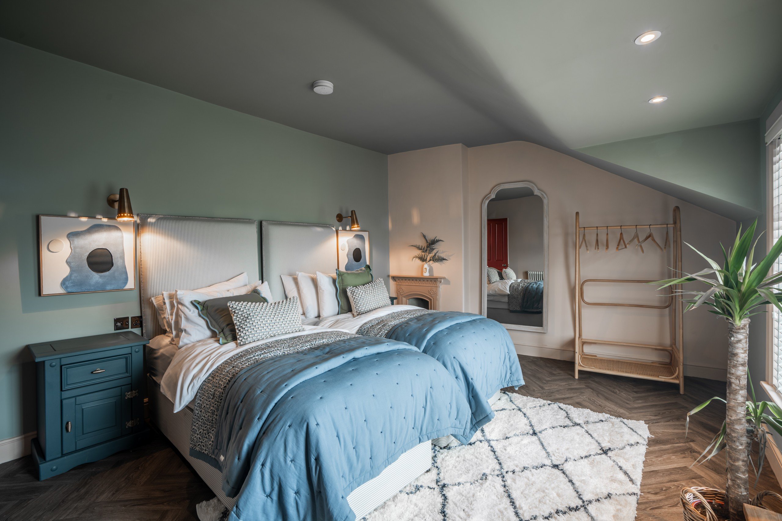 Cozy bedroom with twin beds, blue bedding, and abstract art. Soft lighting, green walls, a large mirror, and a plant accent the space.