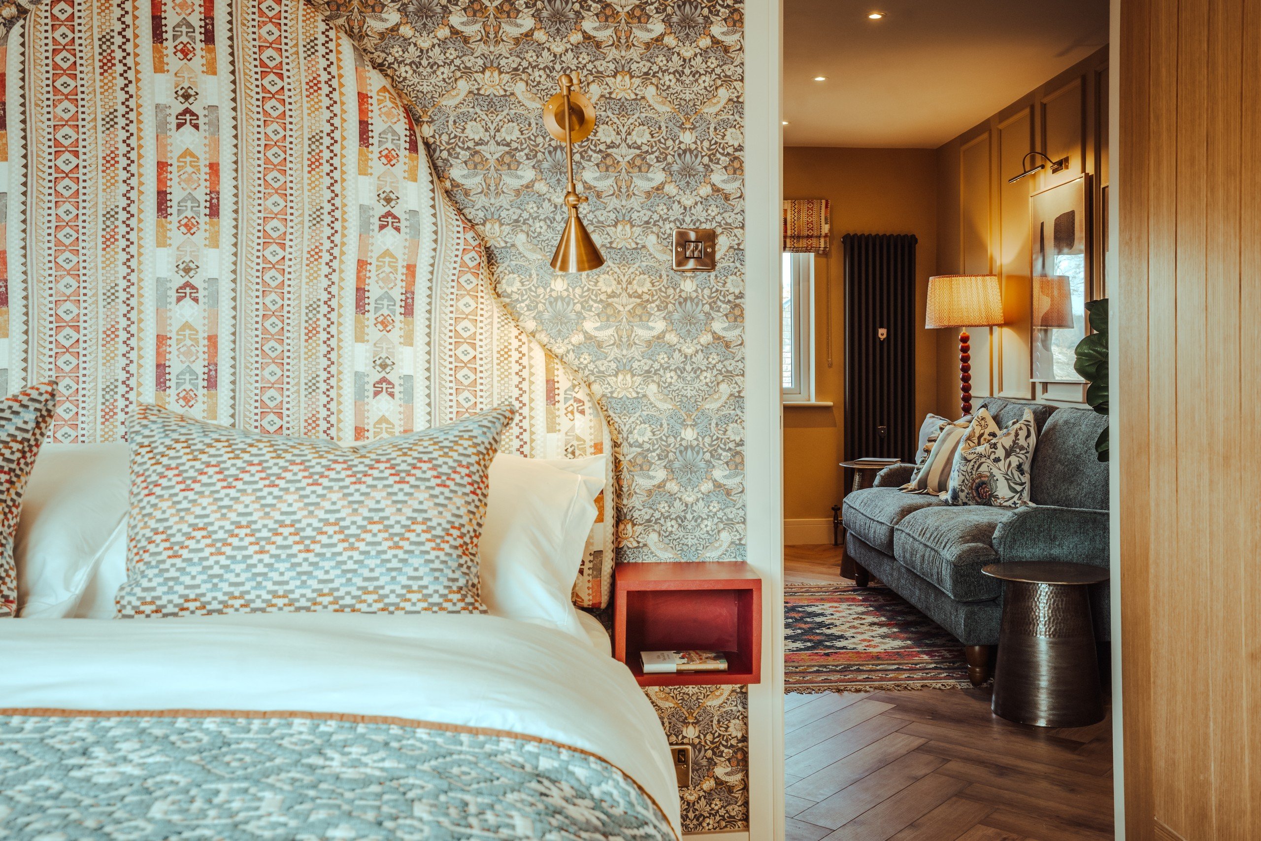Cozy bedroom with patterned wallpaper, colorful pillows, and wooden accents. Bright lamp beside a plush sofa in adjoining room. Warm tones.