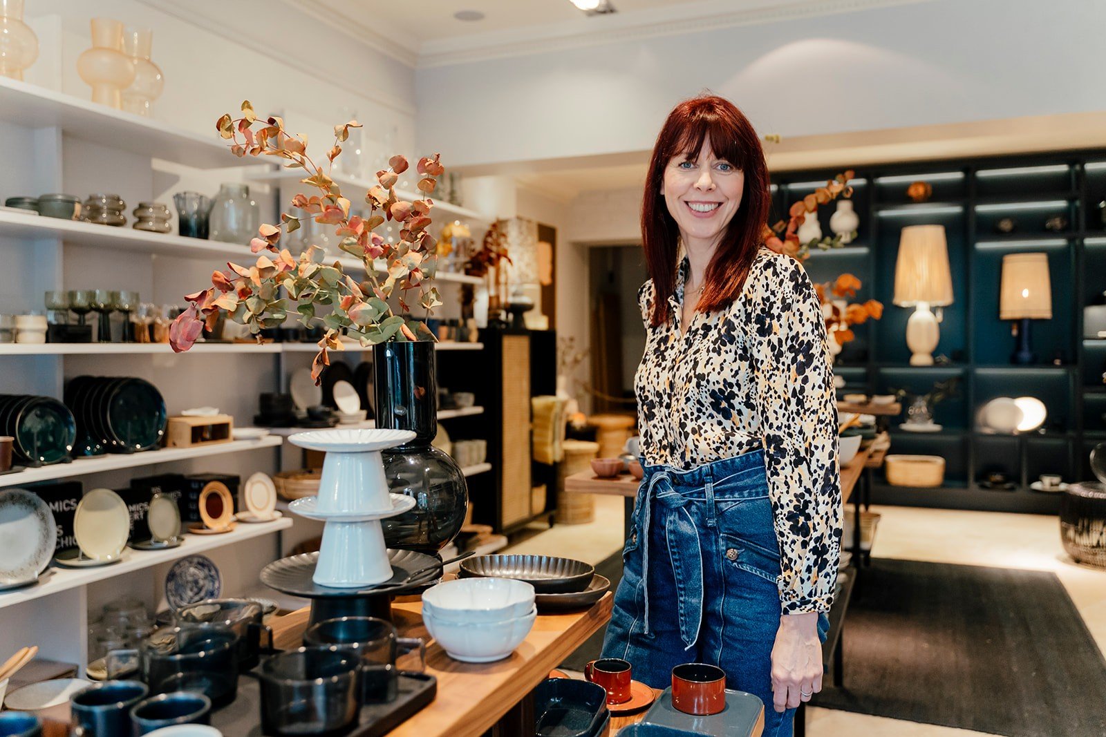 A smiling woman in a patterned blouse stands in a home store surrounded by dishware. Shelves with ceramics and warm lighting are in the background.