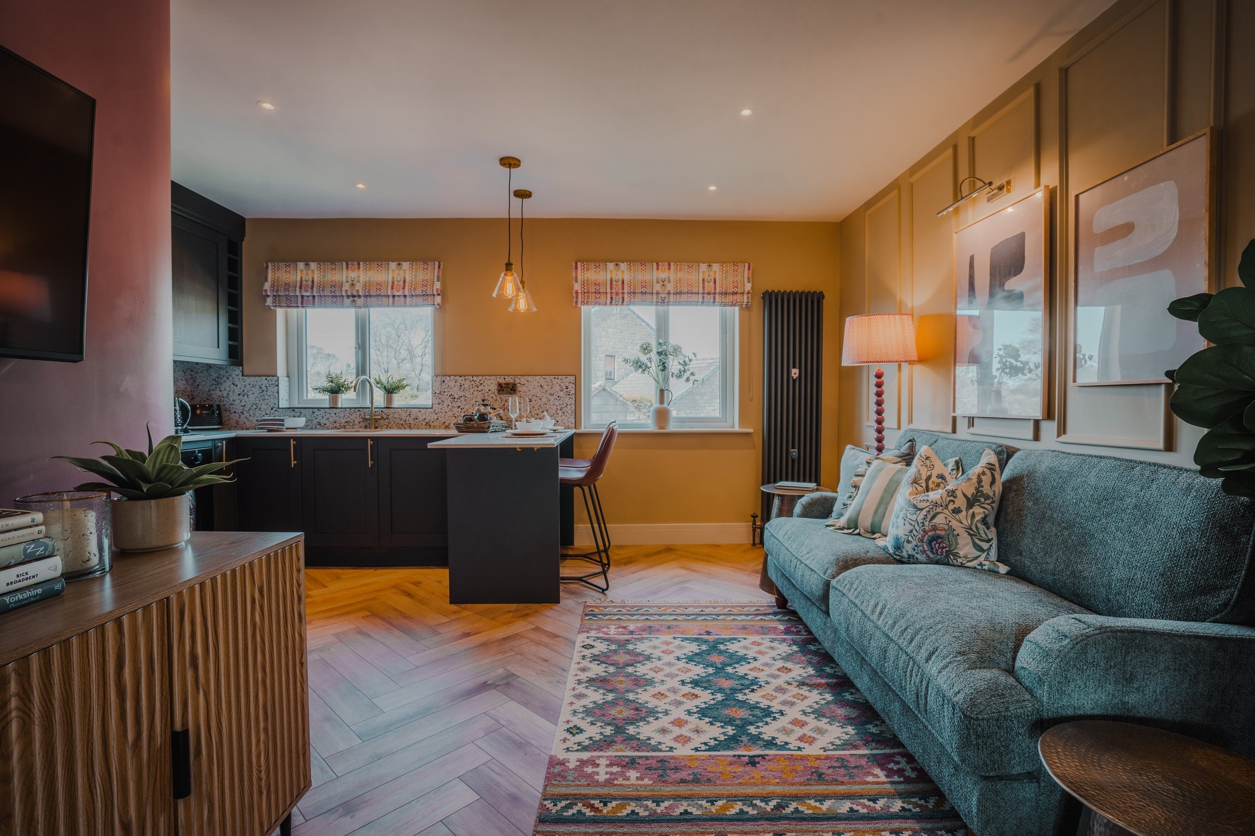 Cozy living room with teal sofa, patterned rug, and warm lighting. Open kitchen with a bar counter, decorative plants, and wall art.