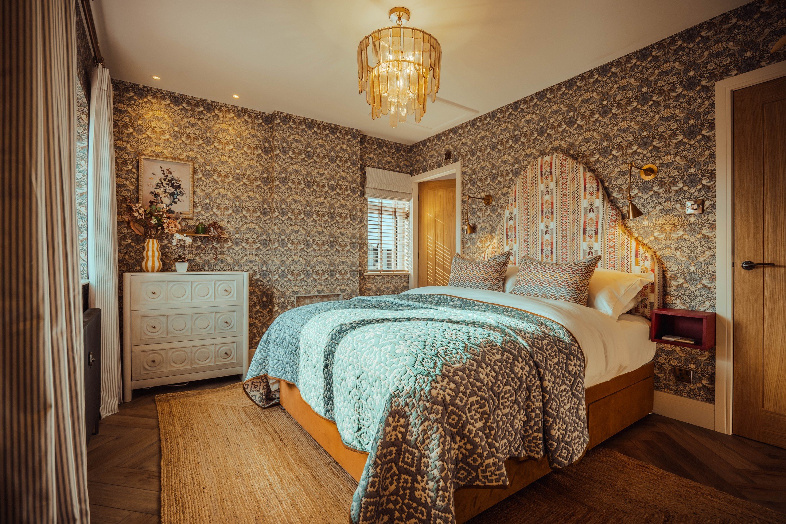 Cozy bedroom with patterned wallpaper, double bed with blue quilt, floral artwork, and a warm chandelier. White dresser and wooden door.