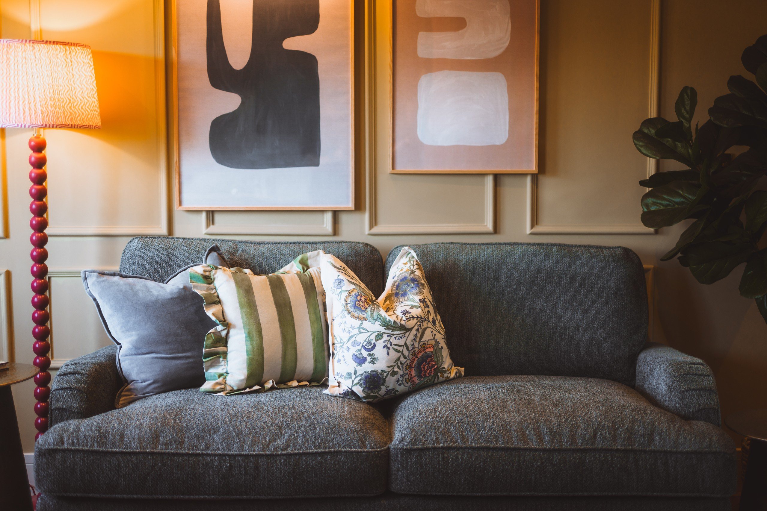 A cozy living room with a gray sofa, colorful patterned pillows, a striped lamp, and abstract art on the wall. Warm, inviting ambiance.