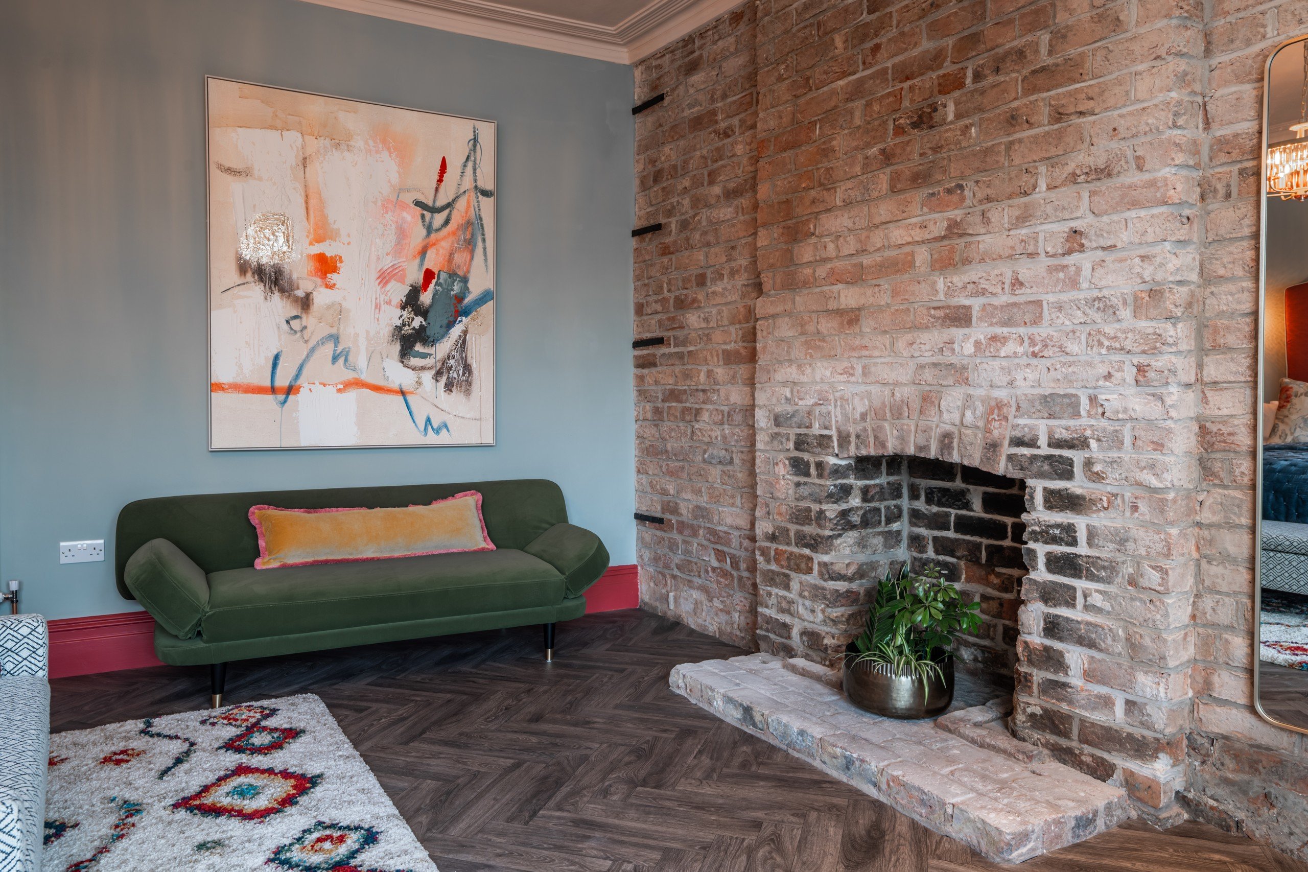 WildKind designed room. Green sofa with an orange pillow under abstract art in a room with blue walls. Exposed brick fireplace, plant, and colourful rug on wood floor.