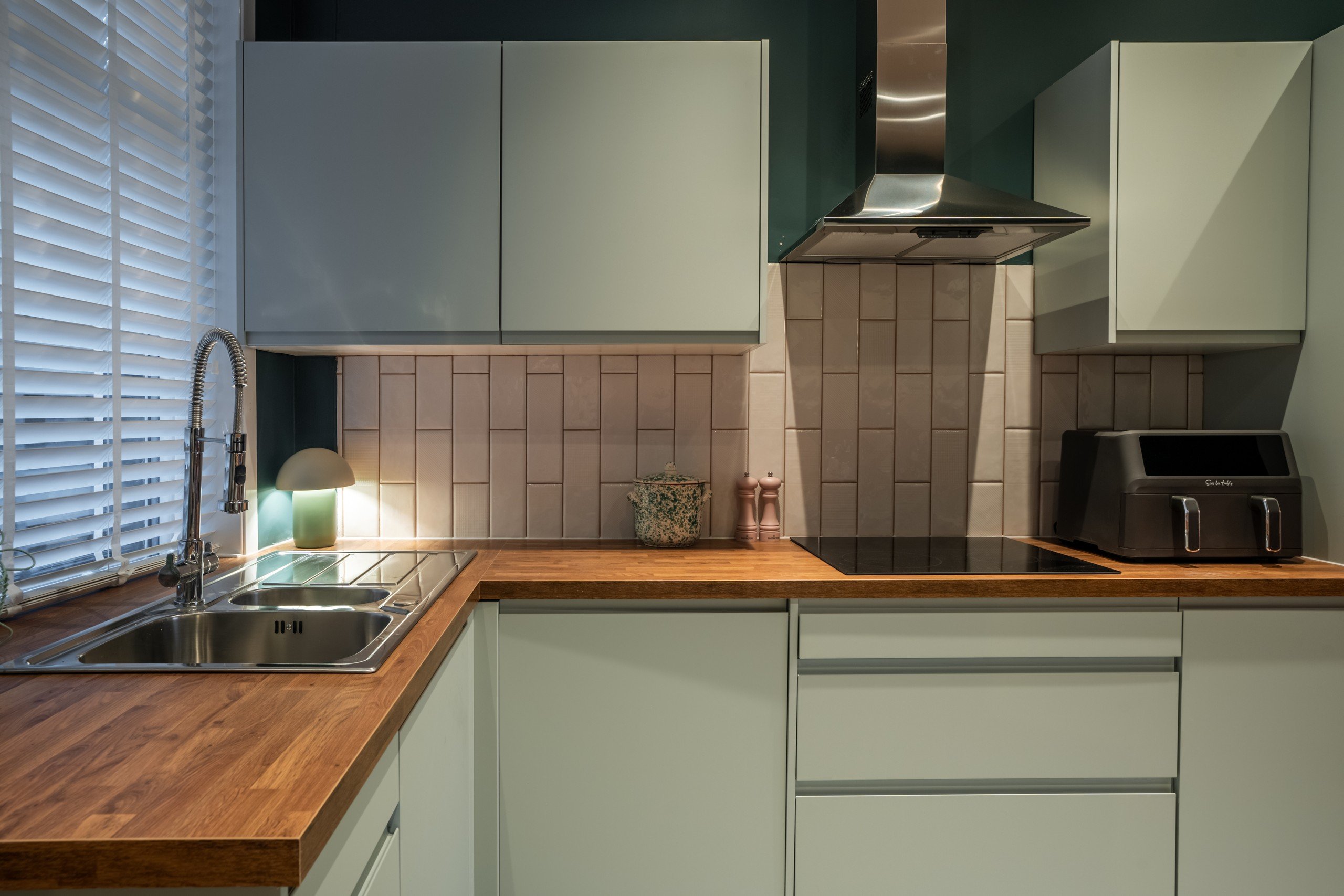 WildKind Designed Modern kitchen with wooden countertops, mint cabinets, and a tiled backsplash. Features a sink, stovetop, toaster, and soft ambient lighting.