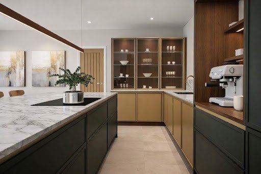 Modern kitchen with marble countertops, green and gold cabinets, and a white coffee machine. Art on walls adds elegance.