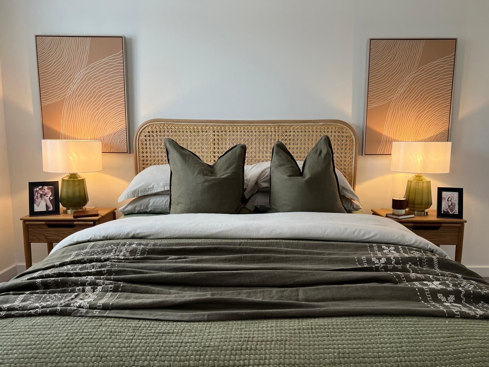 WildKind designed bedroom with rattan headboard, green pillows, and patterned duvet. Two green lamps on wood nightstands, framed photos, and abstract art.