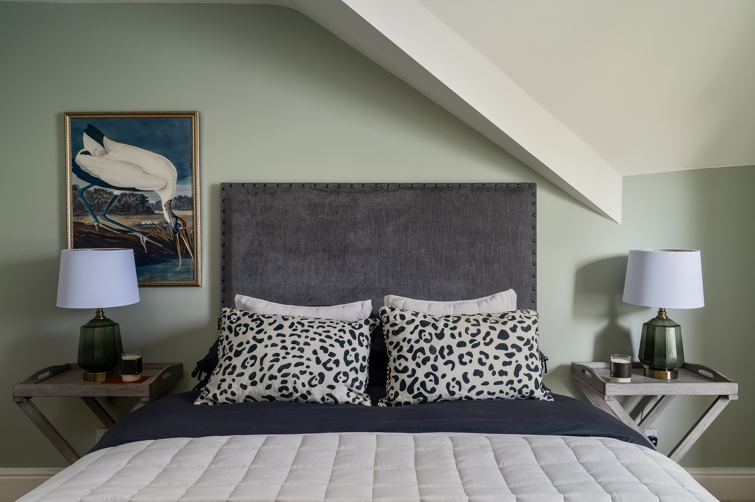 Bed with a grey headboard, leopard print pillows, white quilt, and two green lamps on side tables. A bird painting on a light green wall.