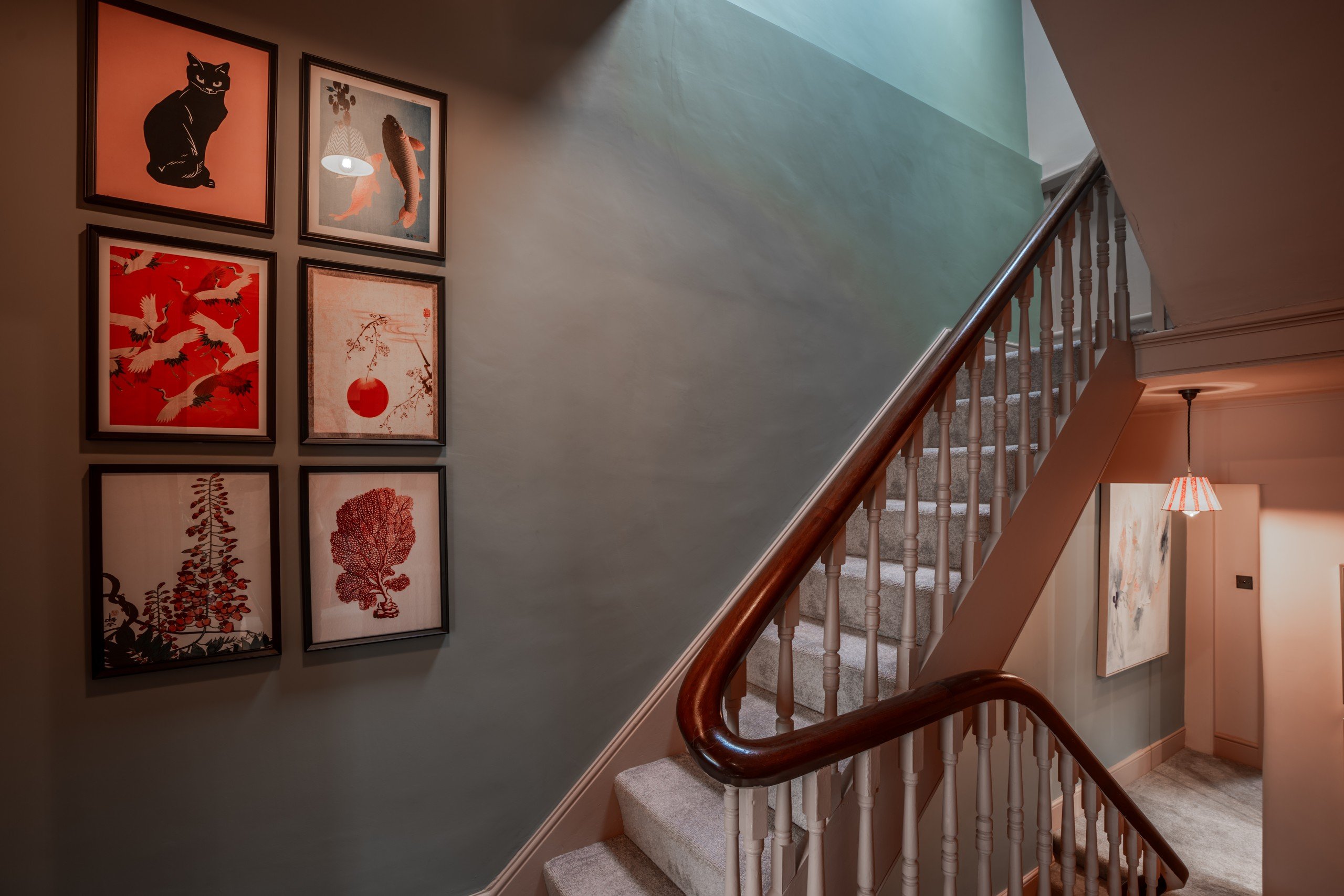 Staircase in a warmly lit hallway with red and black framed art on a green wall. Red striped lampshade emits a cozy glow.