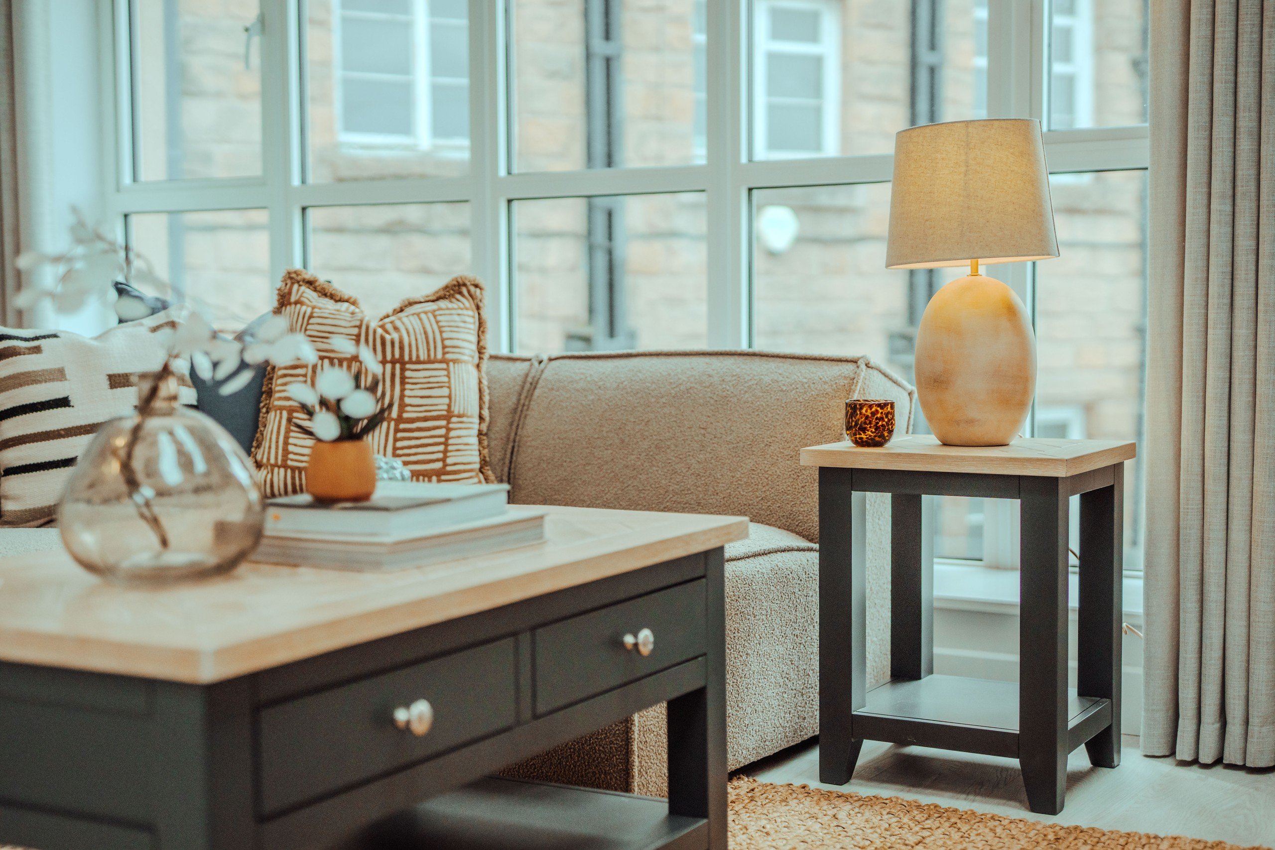 Cozy living room with a beige couch, patterned cushions, a side table with a lamp and cup, and a window with stone building view.
