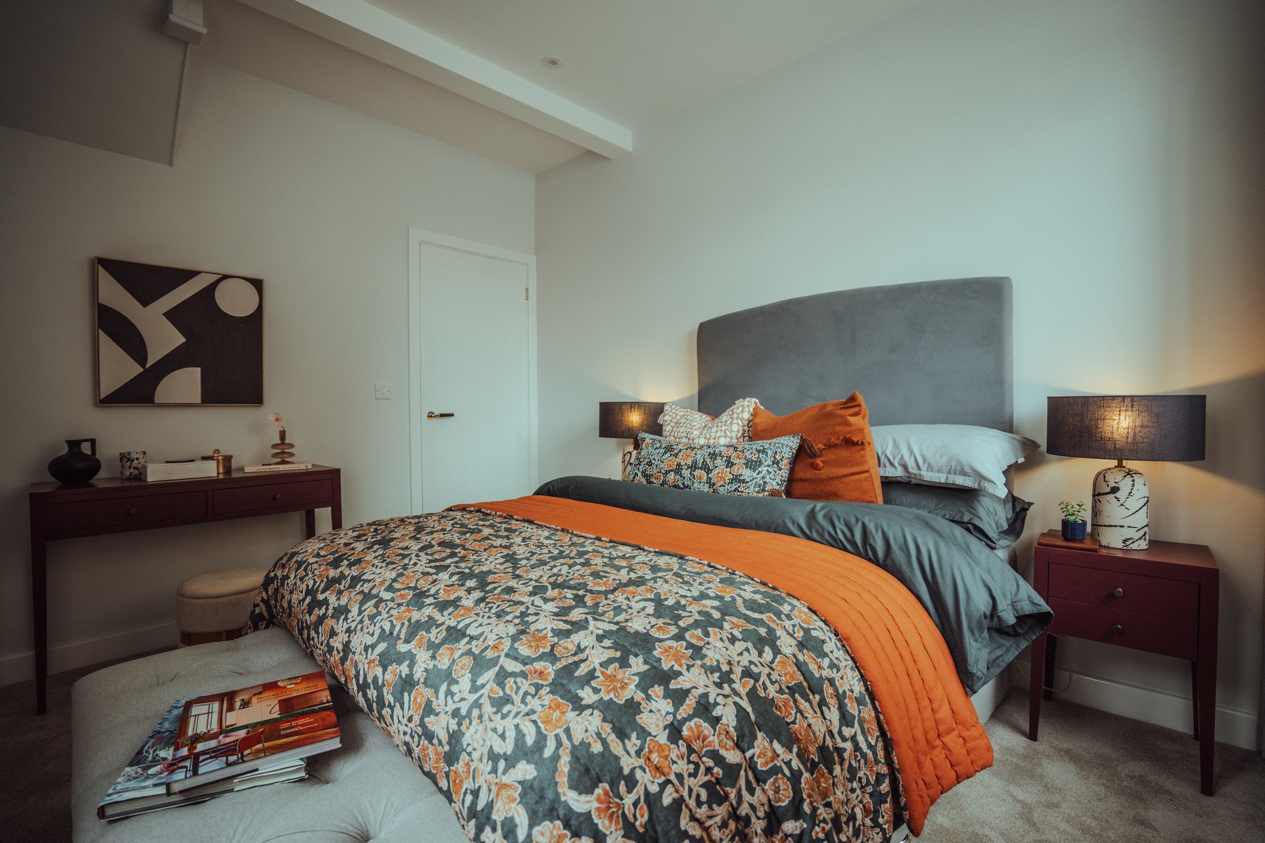 Cozy bedroom with gray bed, orange and floral bedding, and decorative pillows. Two nightstands with lamps, abstract wall art, and a calm ambiance.