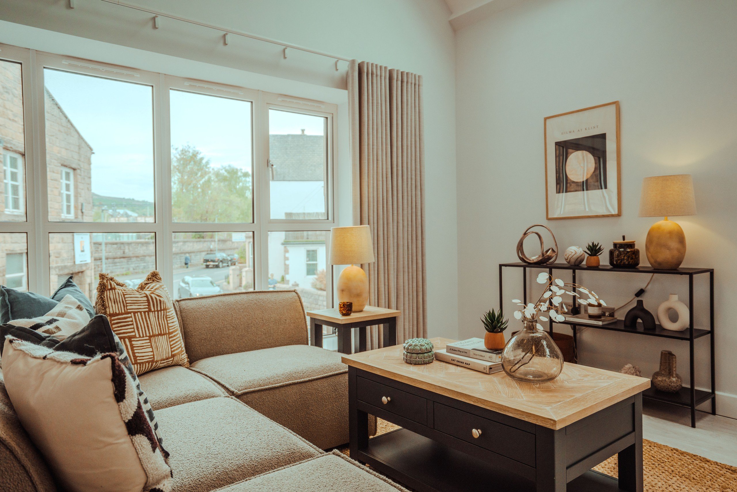 Cozy living room with a beige sofa, patterned pillows, and a wooden coffee table with books and a vase. Large windows, neutral decor.