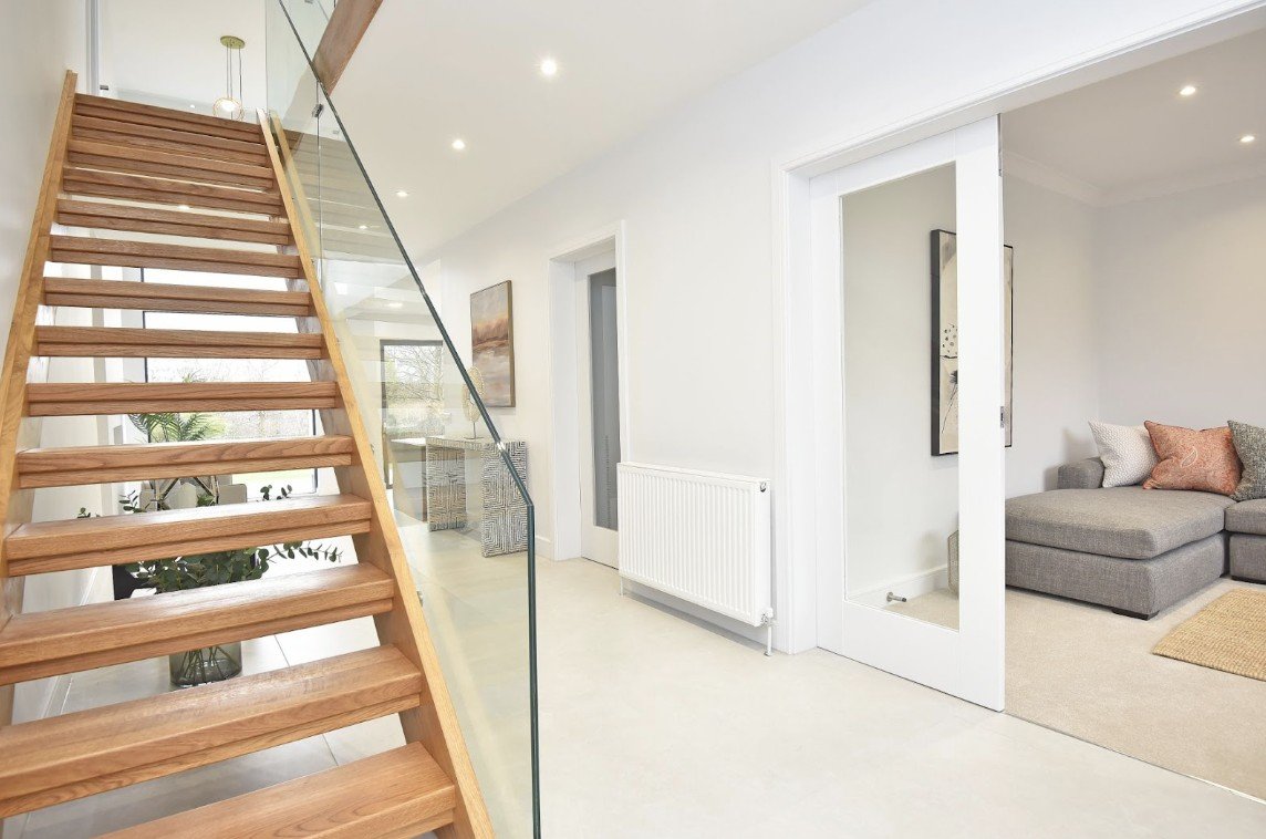 Modern interior with wooden stairs and glass railing, neutral tones. Cozy living area with a gray sofa, white walls, and plants. Bright and inviting.