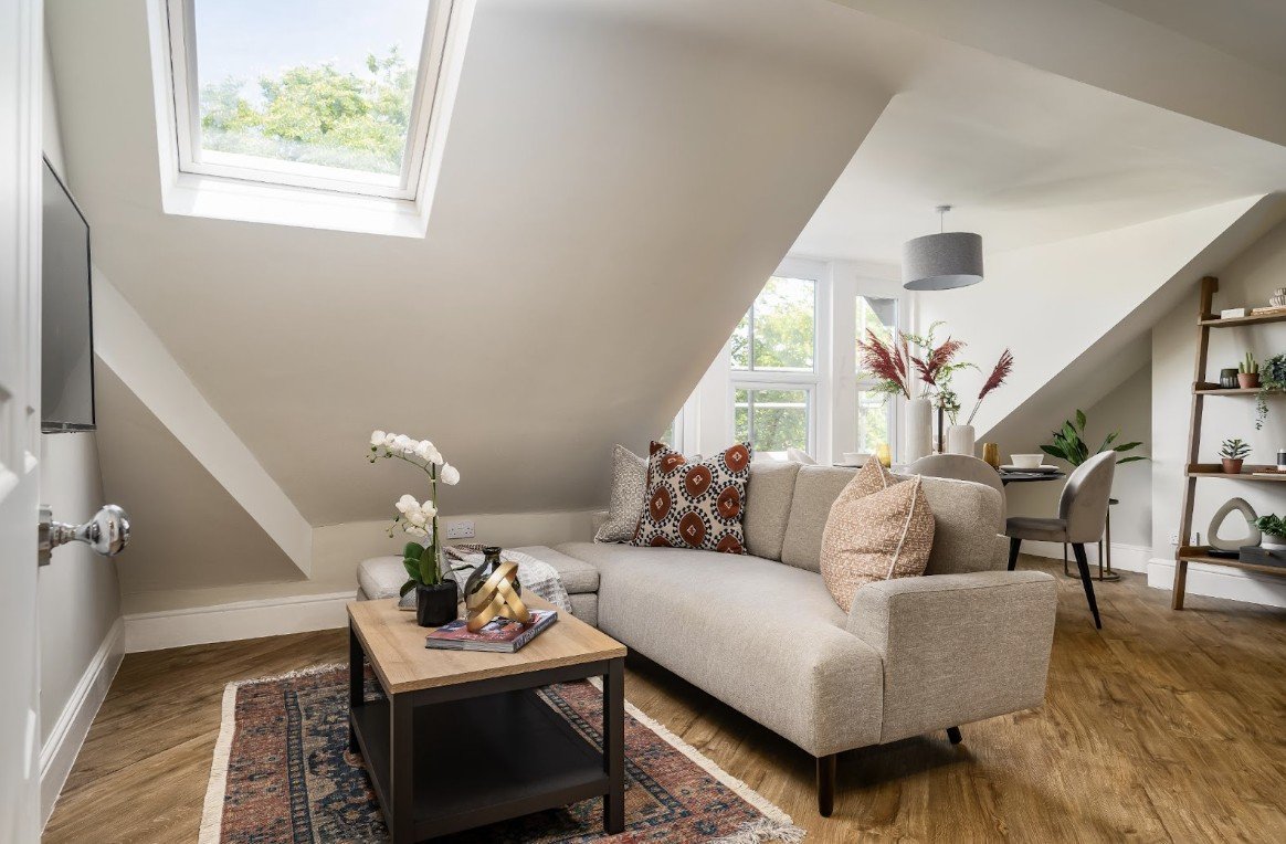 Cozy living room with beige sofa, patterned pillows, coffee table with books and orchid, slanted ceiling, skylight, and wooden floors.