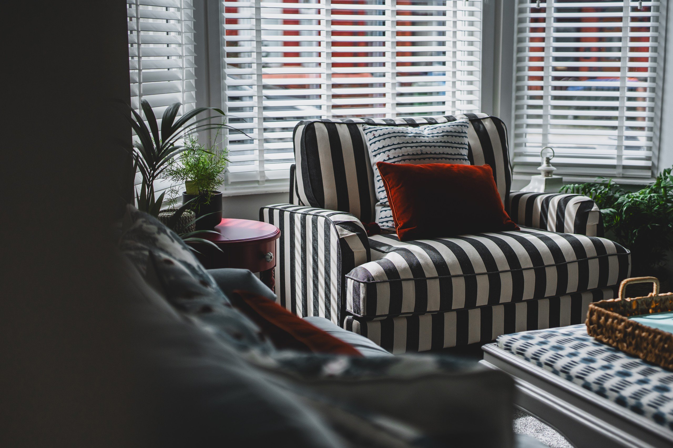 Striped armchair with red pillow in a cozy room. Sunlight filters through blinds, highlighting plants and a red side table. Calm mood.