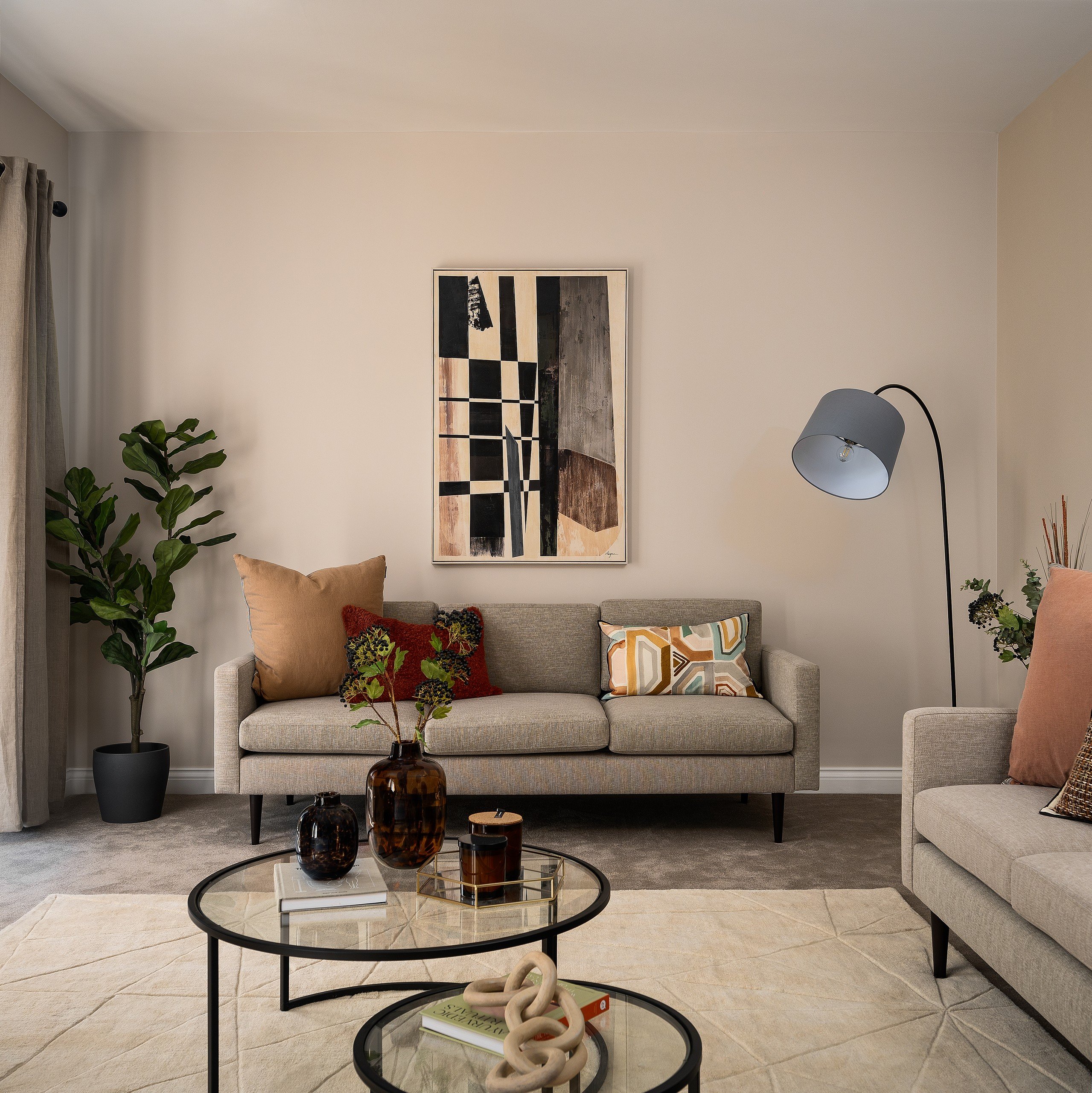 Stylish living room with beige sofas, patterned cushions, abstract art, and a floor lamp. Glass tables hold vases and books. Neutral tones.